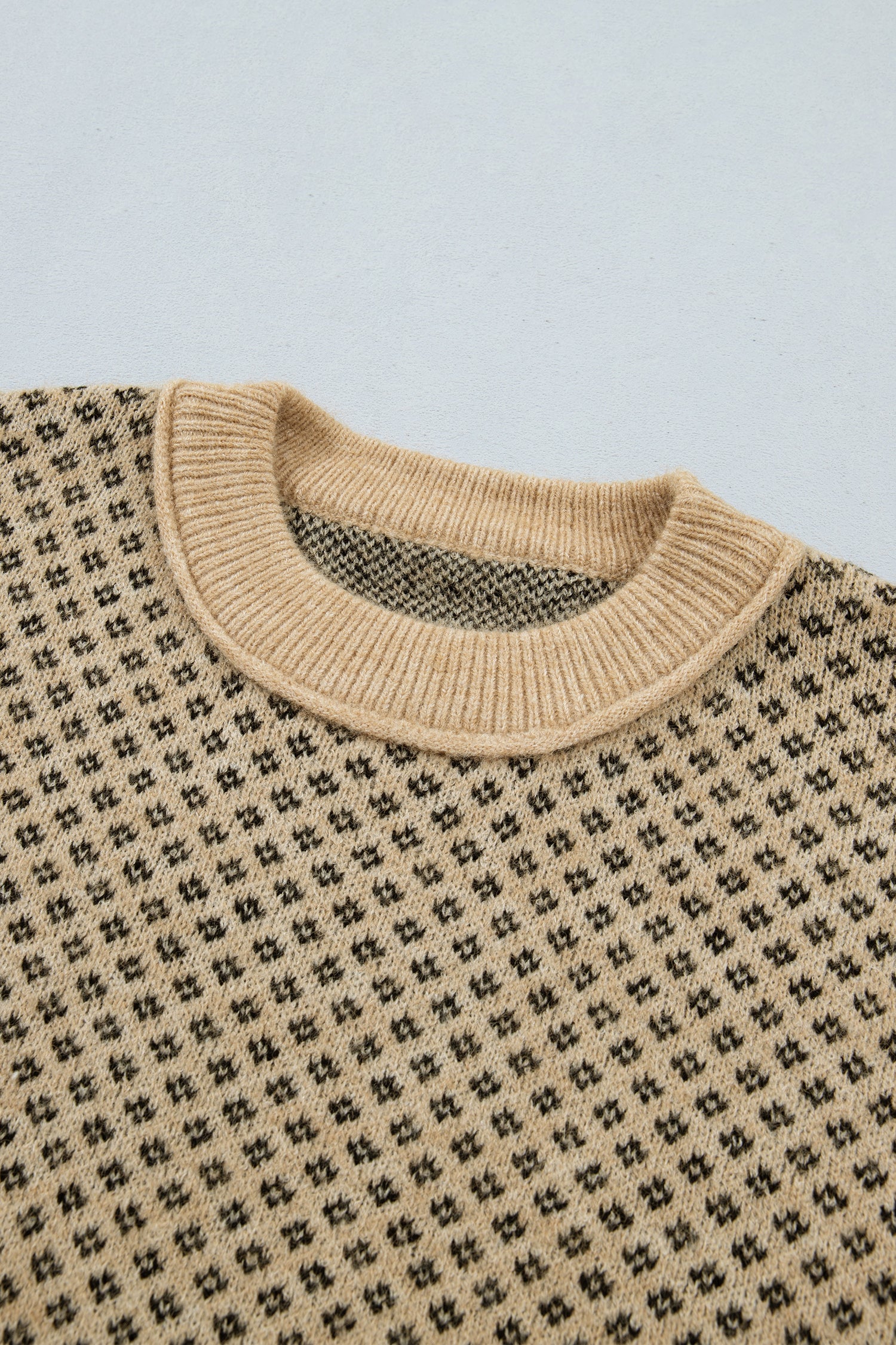 Close-up of a knit crewneck sweater showing the beige ribbed collar and a repeating black geometric pattern on a light beige knit body, set against a light gray background.