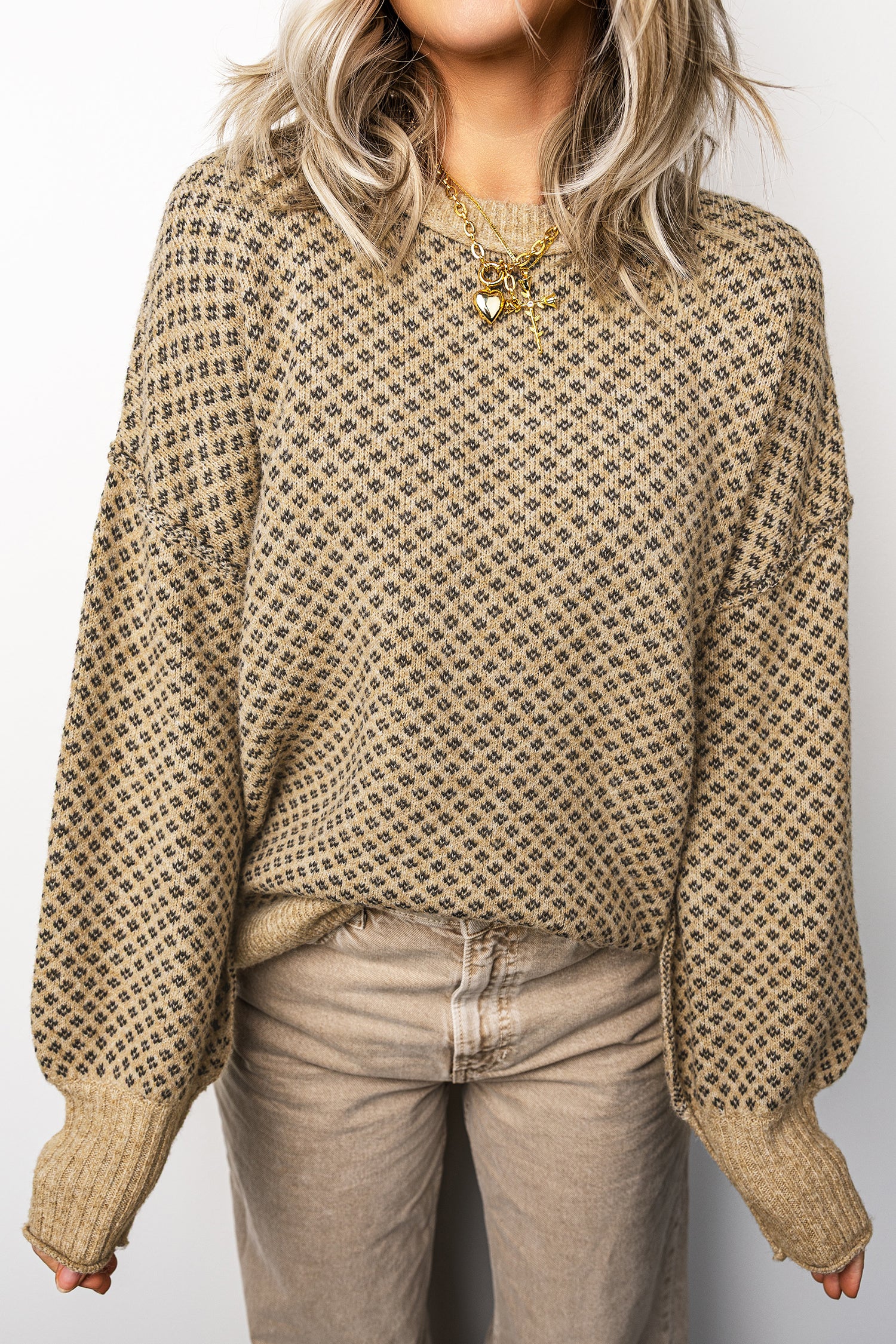 Person wearing a patterned beige sweater and light-colored pants against a plain background
