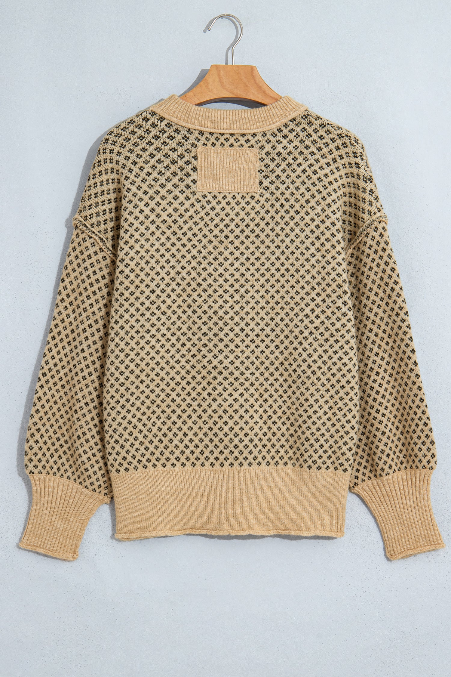 A beige and black knit sweater with a geometric openwork pattern, hanging on a wooden hanger against a light gray background.
