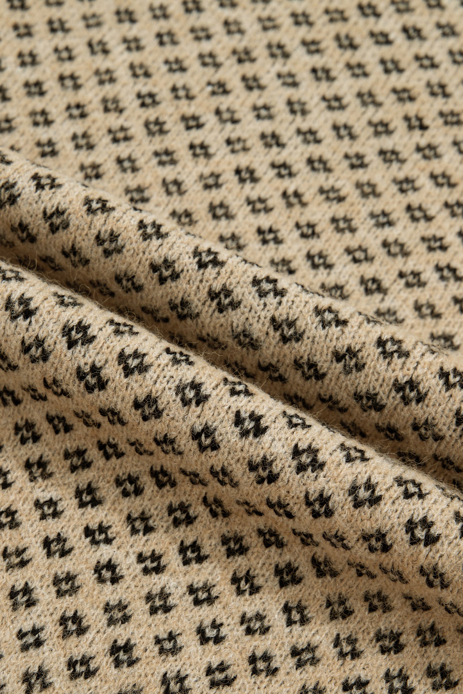Close-up of a knitted fabric with a repeating small black geometric pattern on a warm beige background, shown with a soft fold across the center.