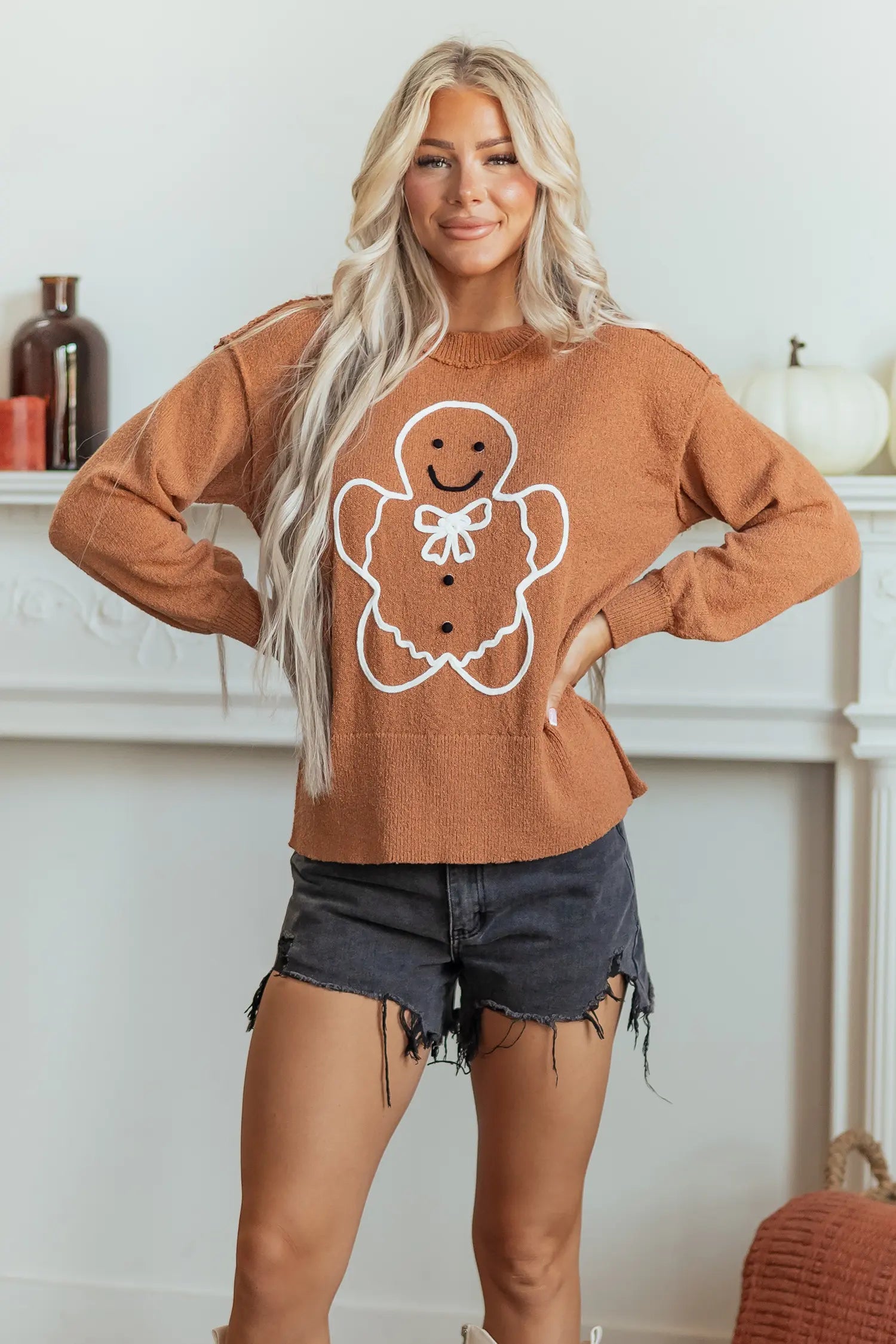 A woman modeling a rust-orange crewneck sweater with a white gingerbread man graphic on the front, paired with dark denim shorts, standing in front of a white fireplace mantel.