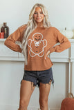 A woman modeling a rust-orange crewneck sweater with a white gingerbread man graphic on the front, paired with dark denim shorts, standing in front of a white fireplace mantel.