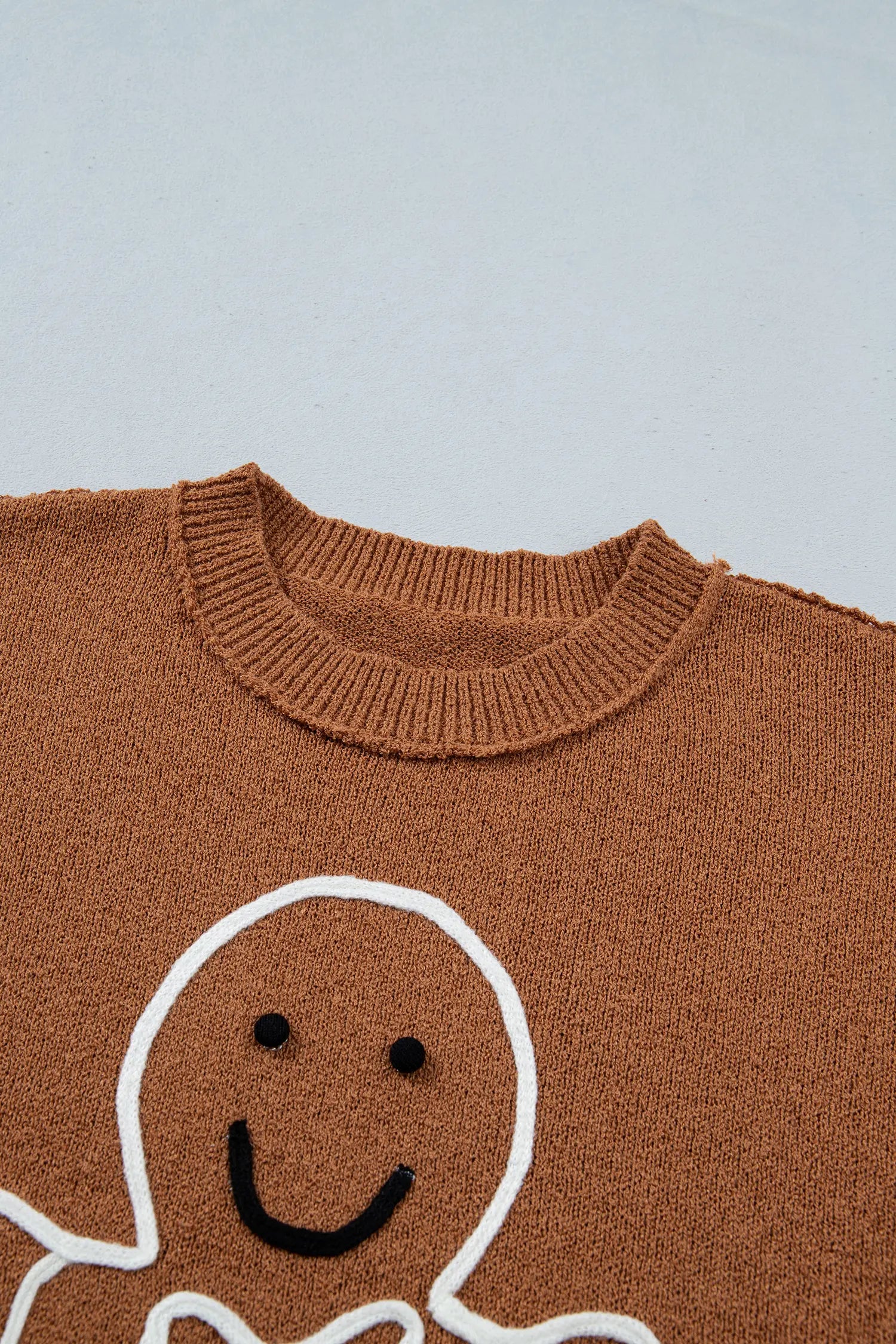 Close-up of a brown knit sweater featuring a white and black gingerbread man face graphic on the front, set against a light gray background.