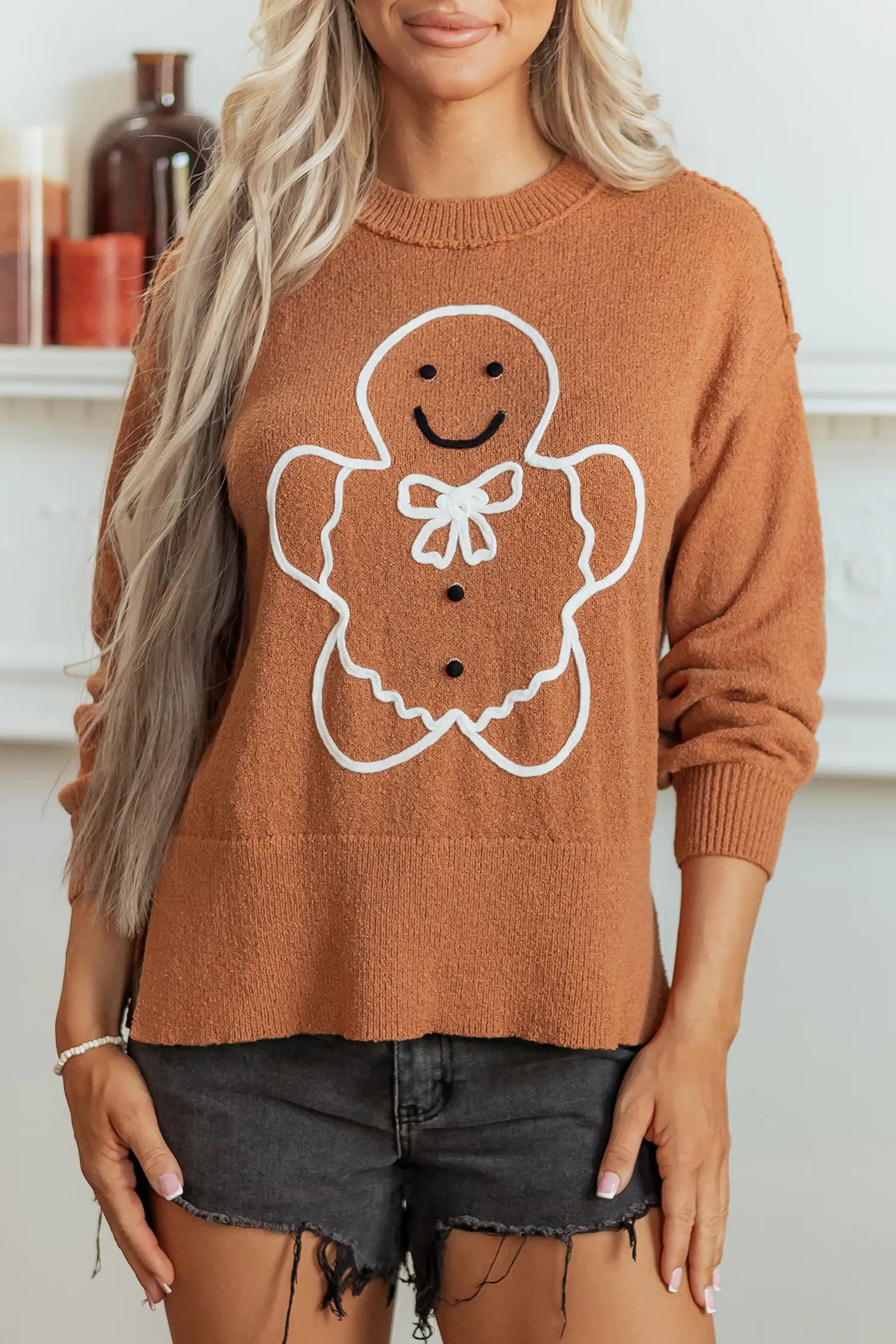 A woman wearing a brown crewneck sweater with a white outline of a gingerbread man on the front, paired with dark gray denim shorts.