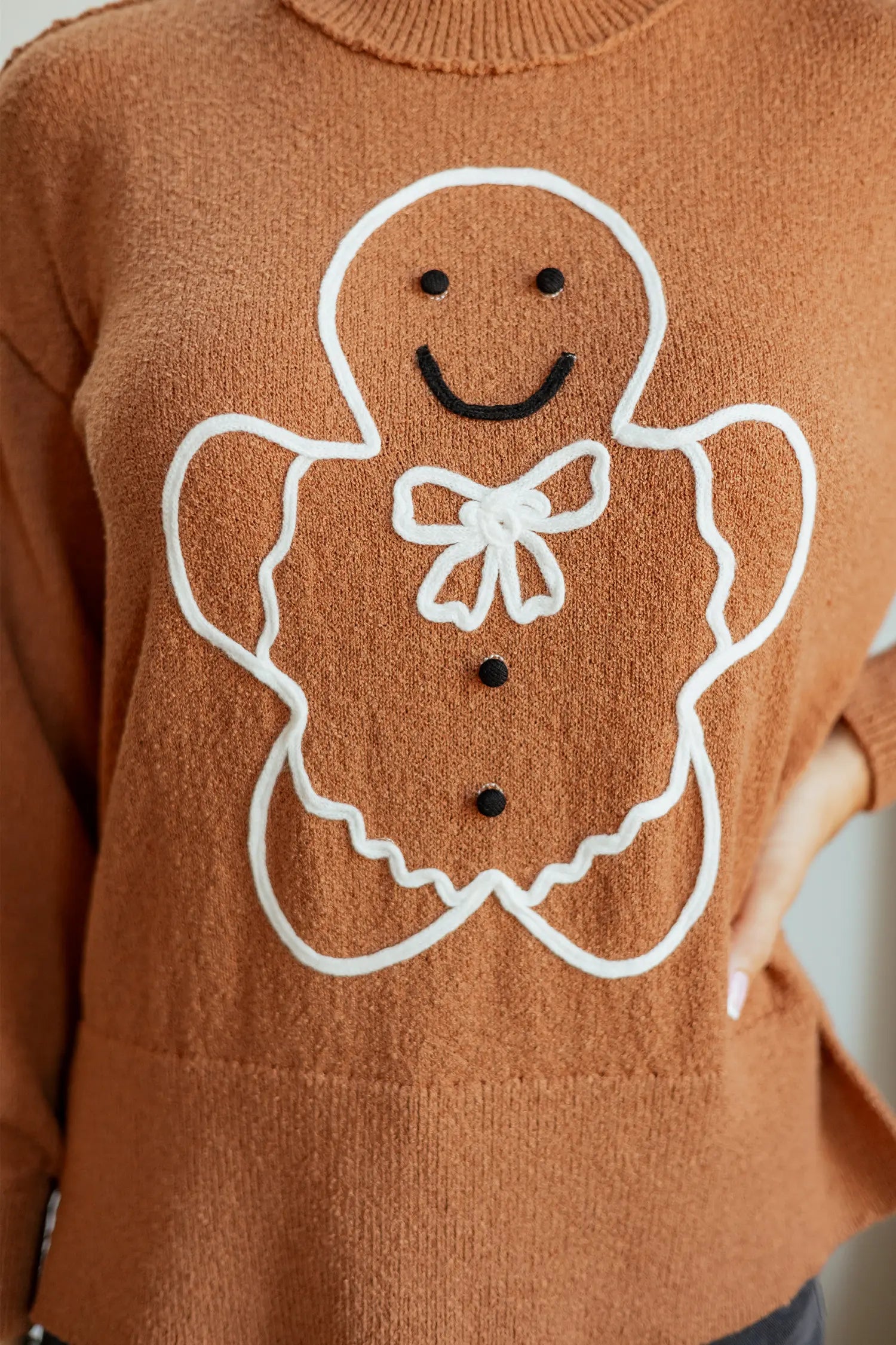 A close-up of a brown knit sweater featuring a large white gingerbread man outline with black facial features and buttons on the front.