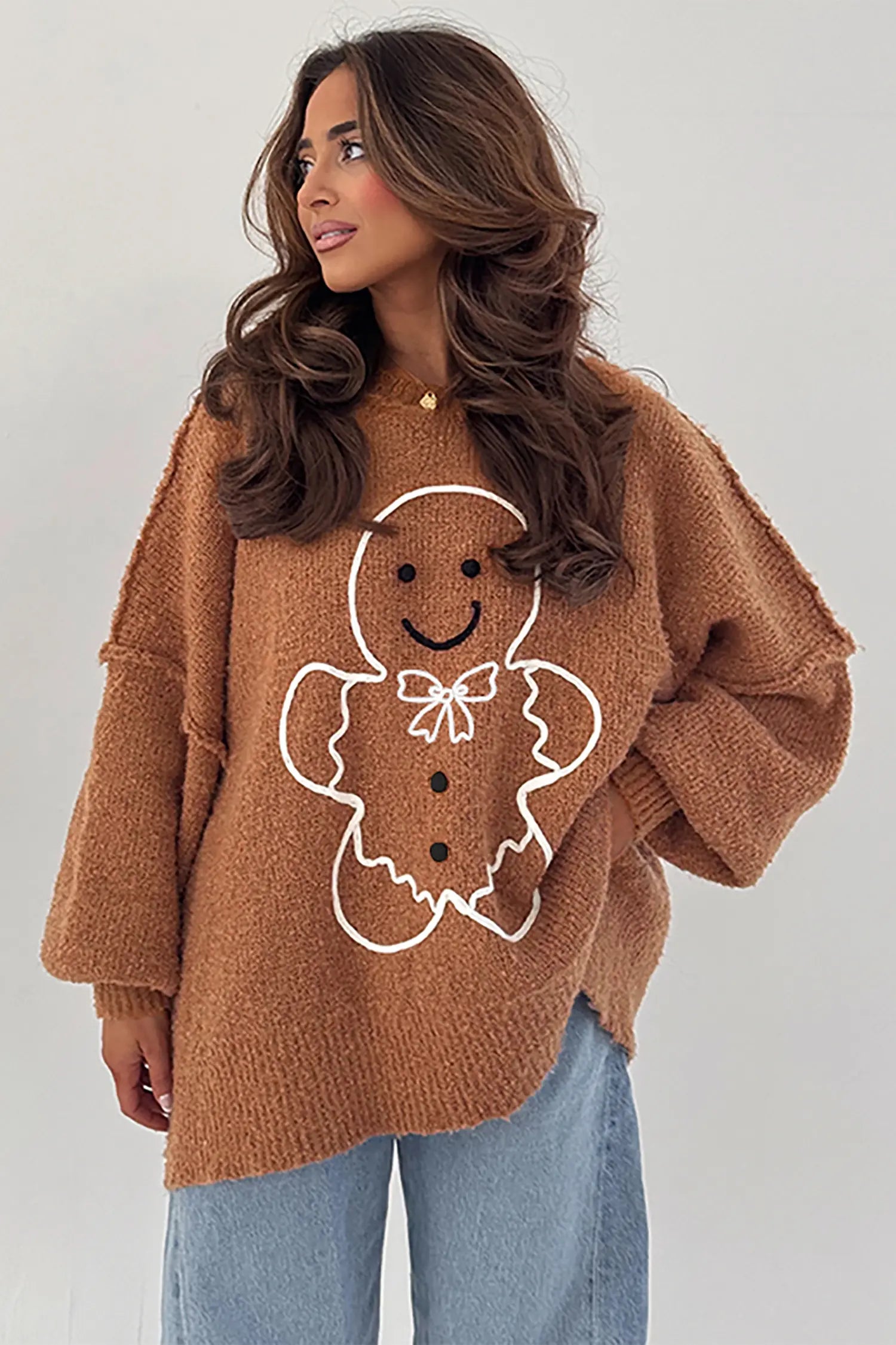 A woman wearing a brown fuzzy knit sweater with a white gingerbread man graphic on the front, paired with light blue jeans, against a plain light gray background.