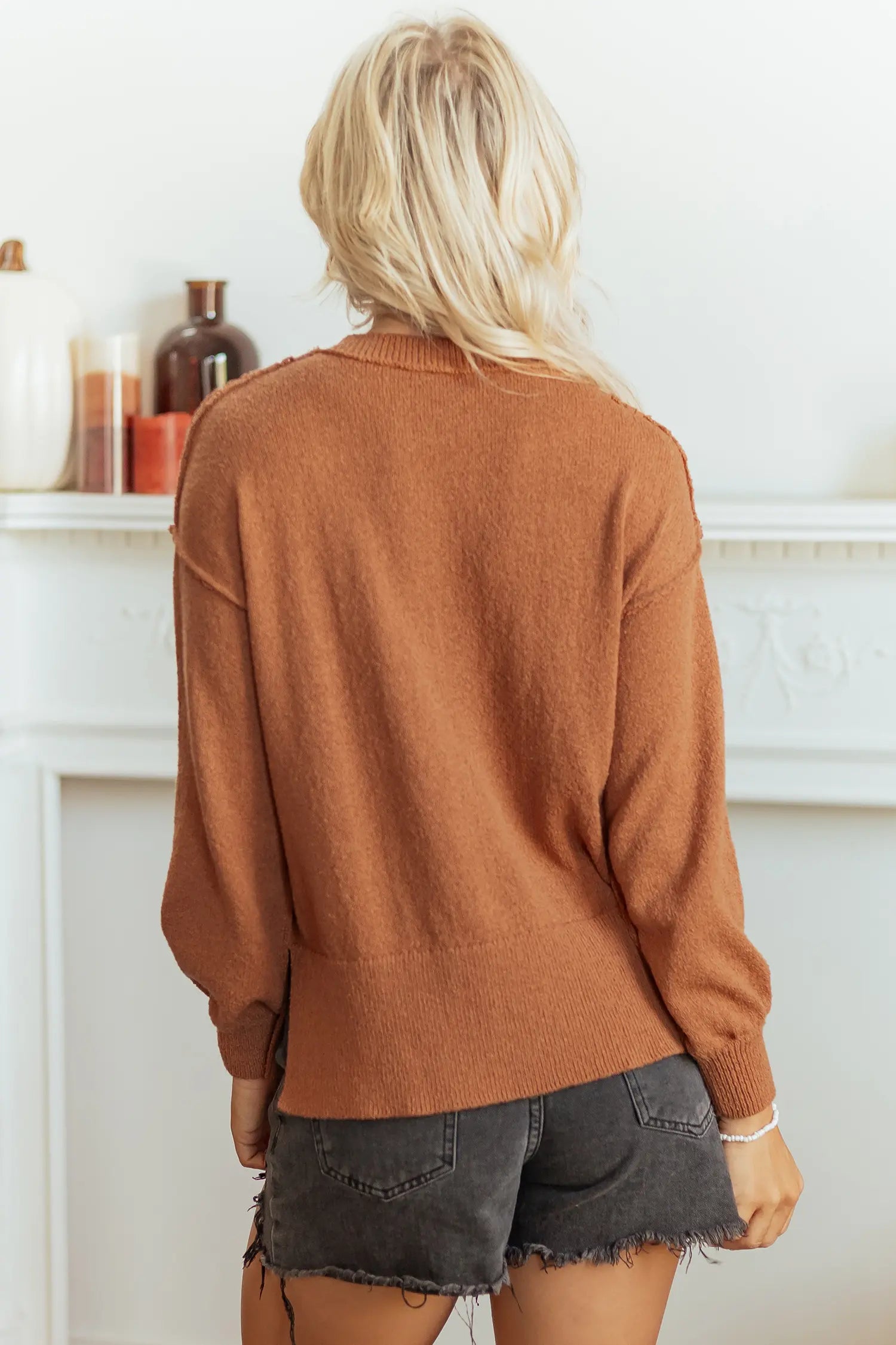 A woman viewed from behind wearing a rust-brown knit cardigan and dark gray denim shorts in front of a white fireplace mantel.