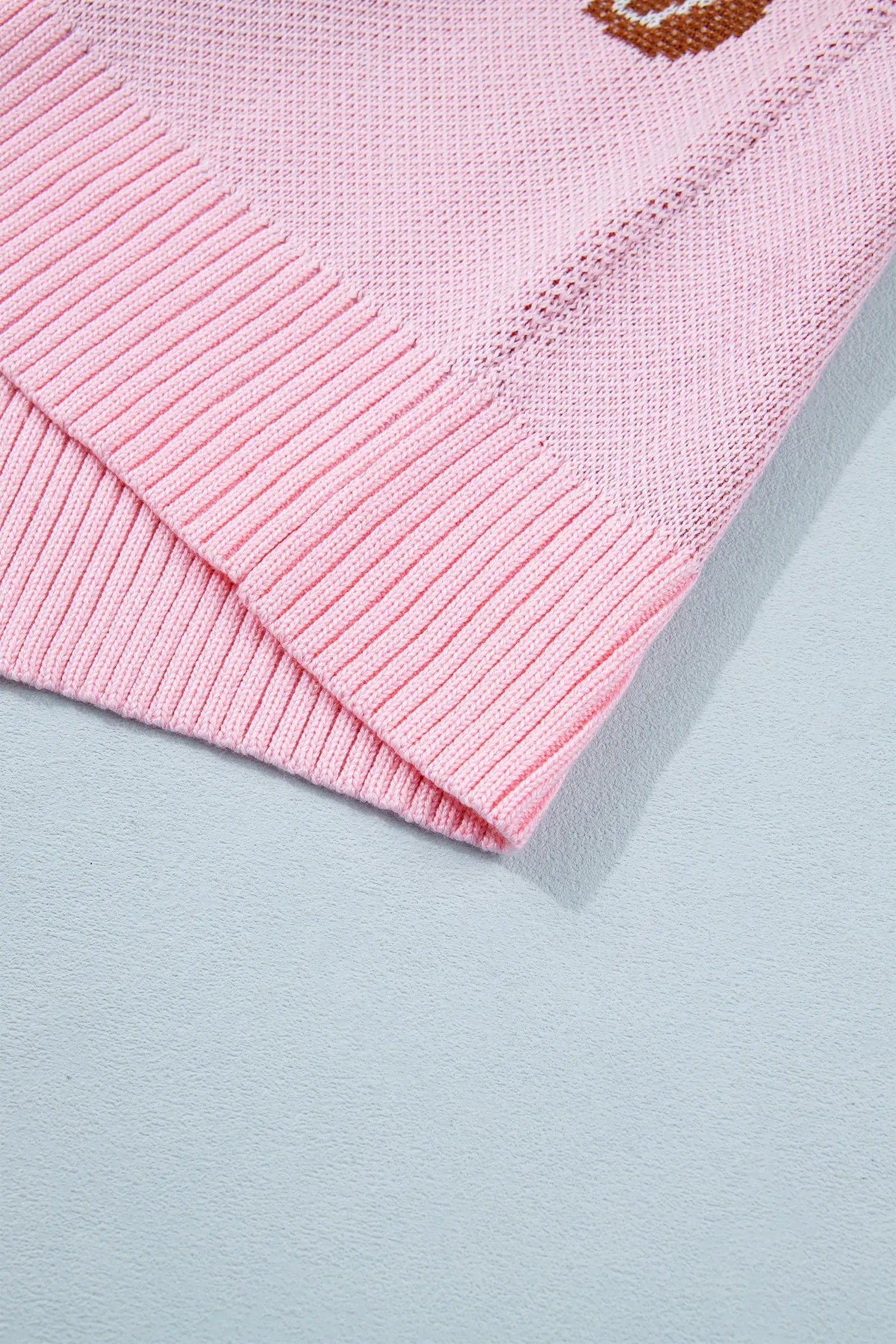 Close-up of light pink knit garment showing ribbed cuff and brown embroidered letter on pale blue.