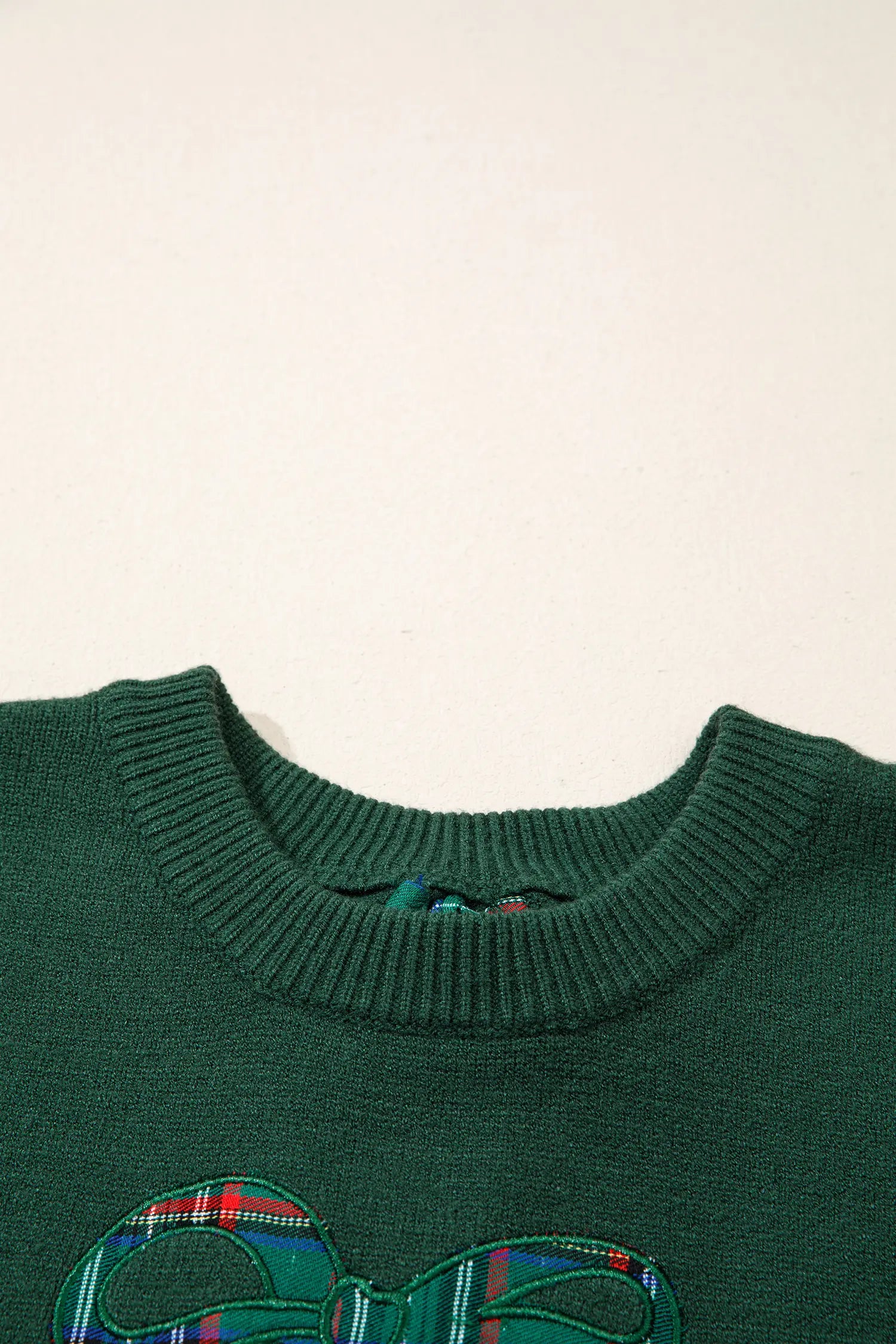 Close-up of dark green sweater with ribbed crew neck and plaid bow graphic.
