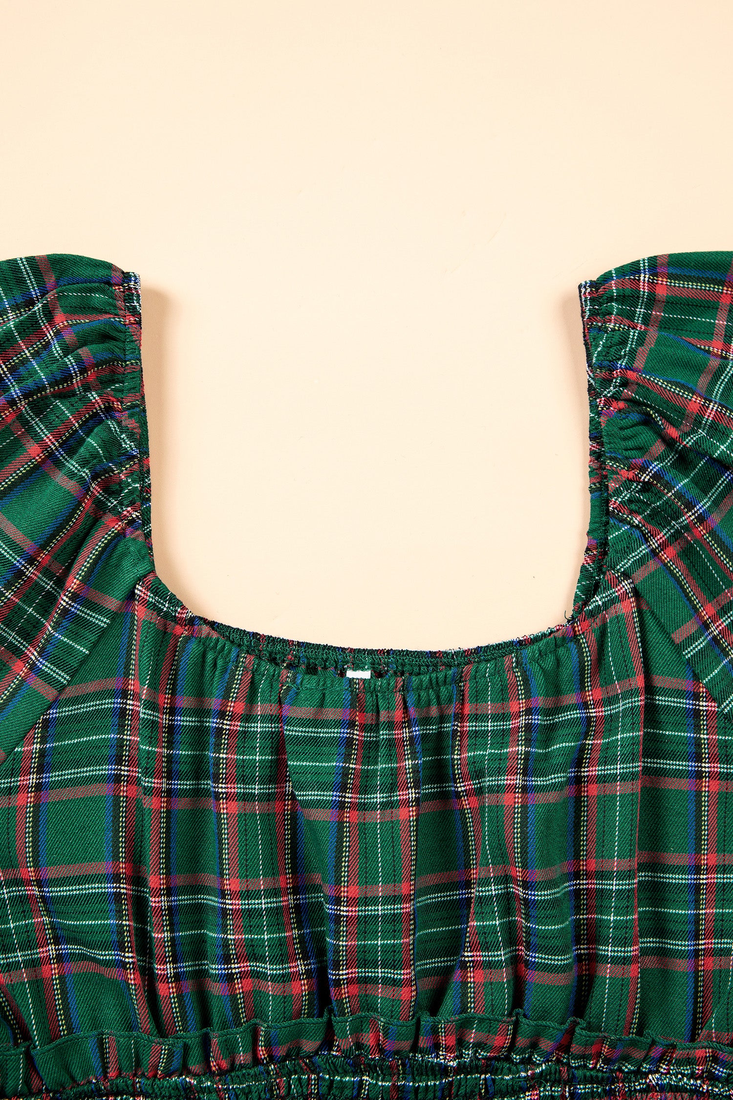 A close-up of a green tartan plaid top with ruffled shoulder straps and a smocked bodice, shown against a solid pale peach background.