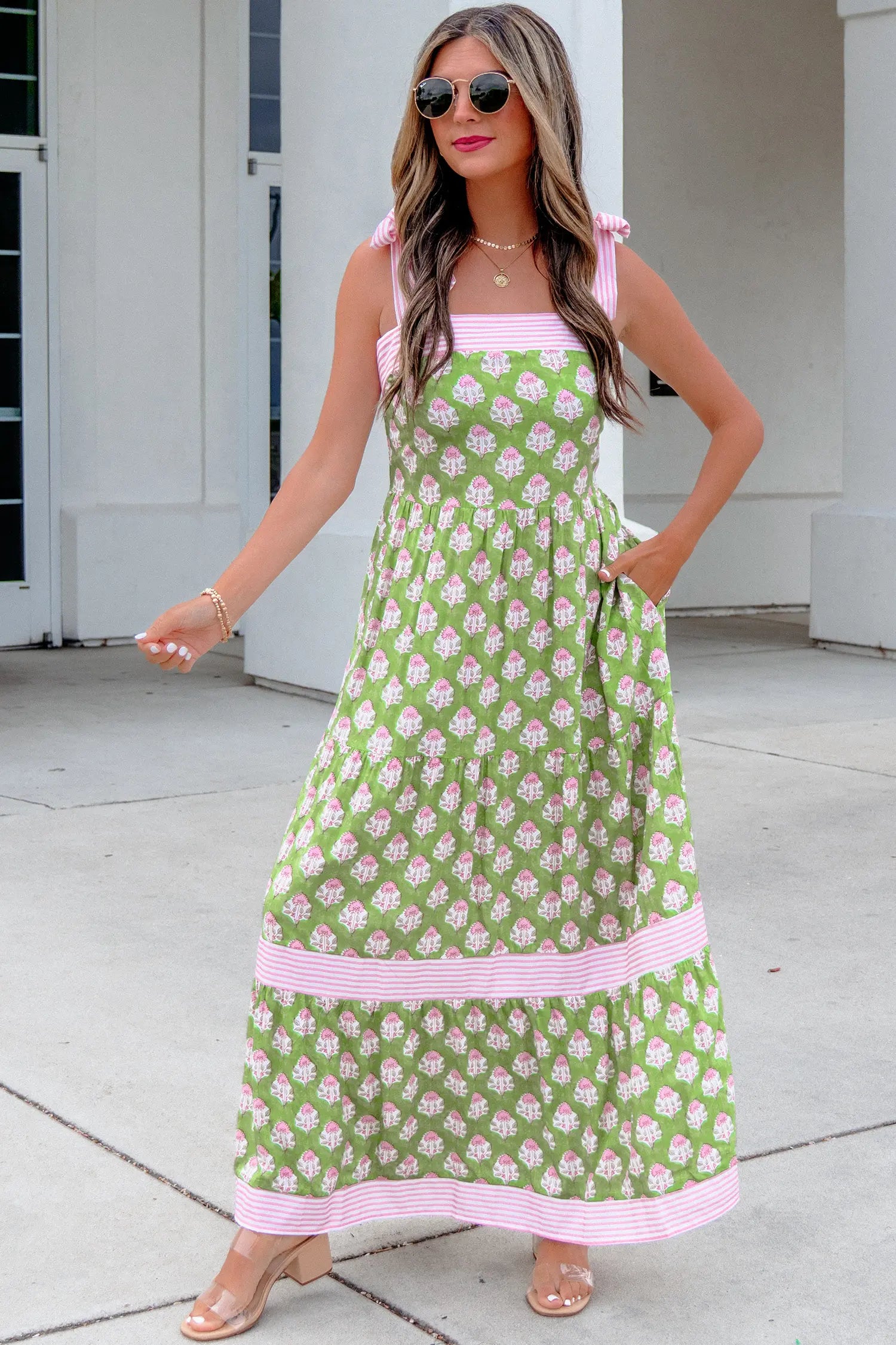 A woman modeling a sleeveless green maxi dress with a pink and white floral pattern, pink gingham trim, and tie straps, standing outdoors on a concrete walkway.