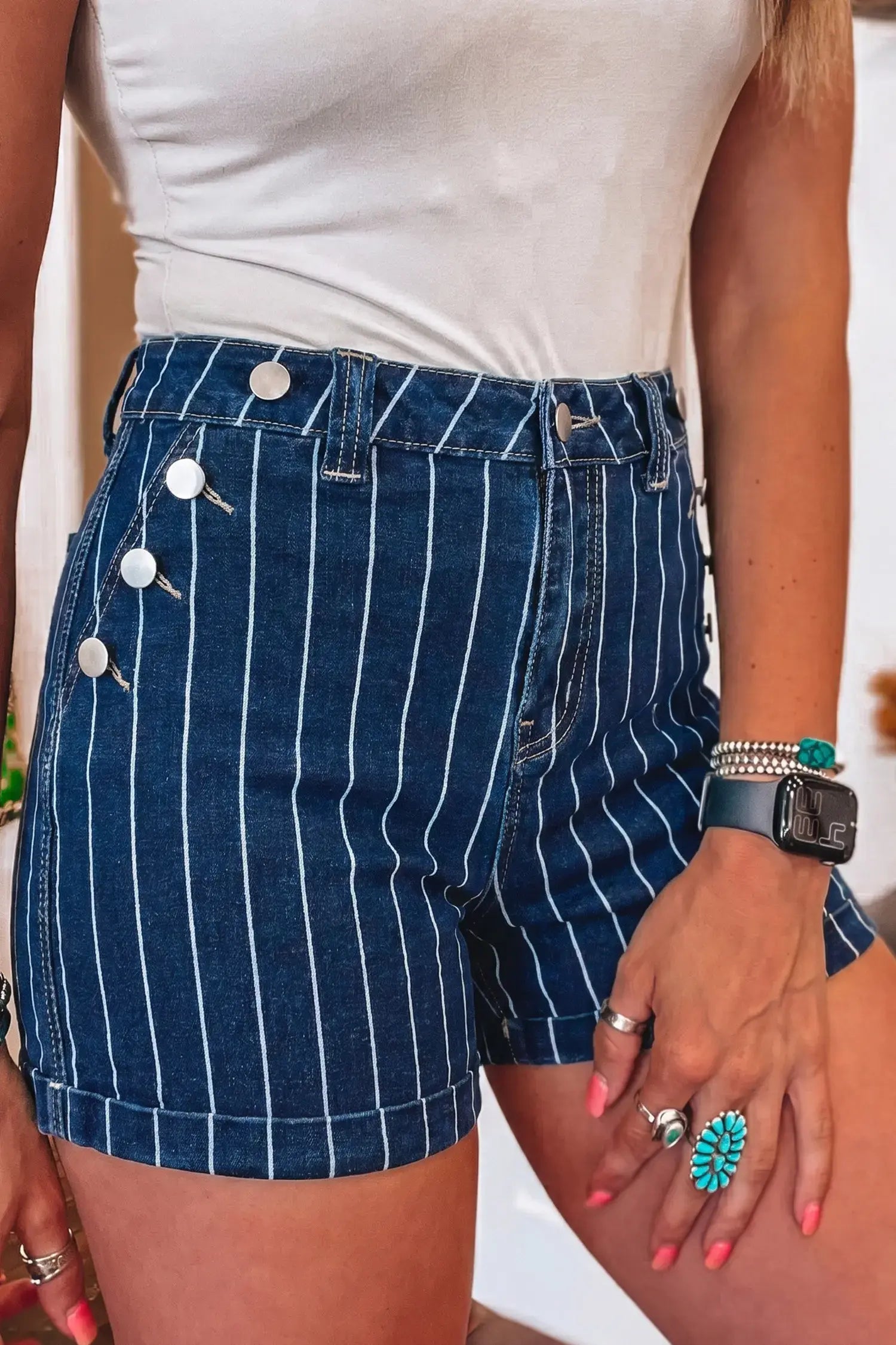 A close-up of a person wearing dark blue denim shorts with thin white vertical pinstripes and decorative silver-tone buttons on the side, paired with a white top.