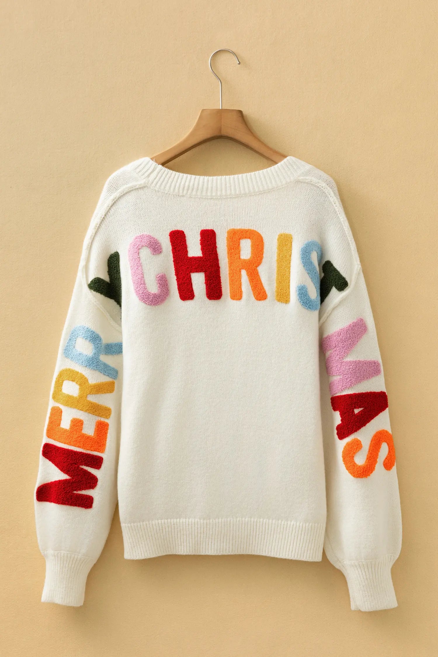 A cream-colored knit sweater hanging on a wooden hanger against a warm beige background. The back of the sweater features colorful, raised lettering spelling 'CHRIST' across the shoulders, with 'MERRY' on one sleeve and 'VAS' on the other, in pink, orange, blue, and green.