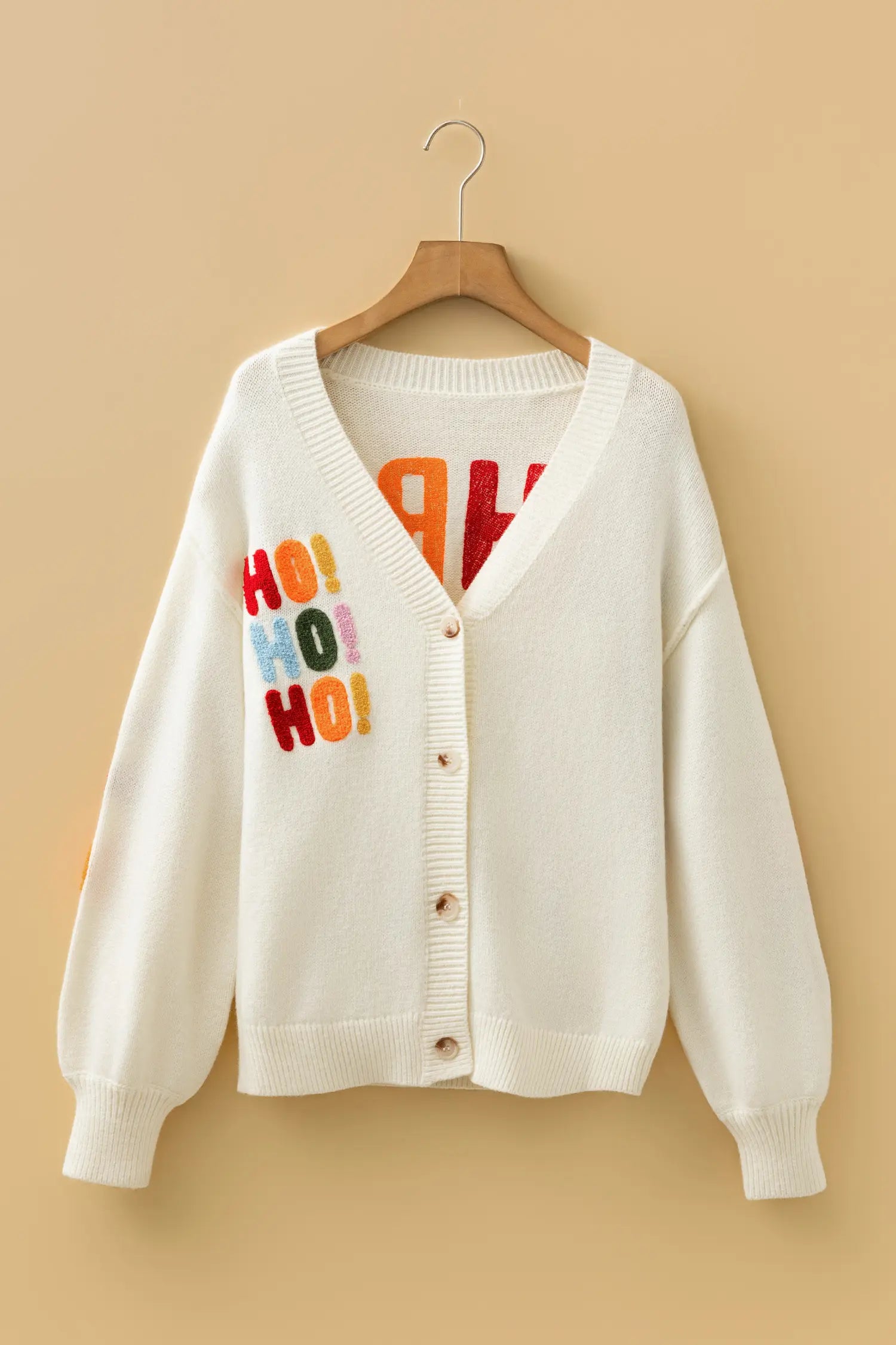 An off-white knit cardigan sweater with colorful 'HO! HO! HO!' text on the left chest, hanging on a wooden hanger against a warm beige background.