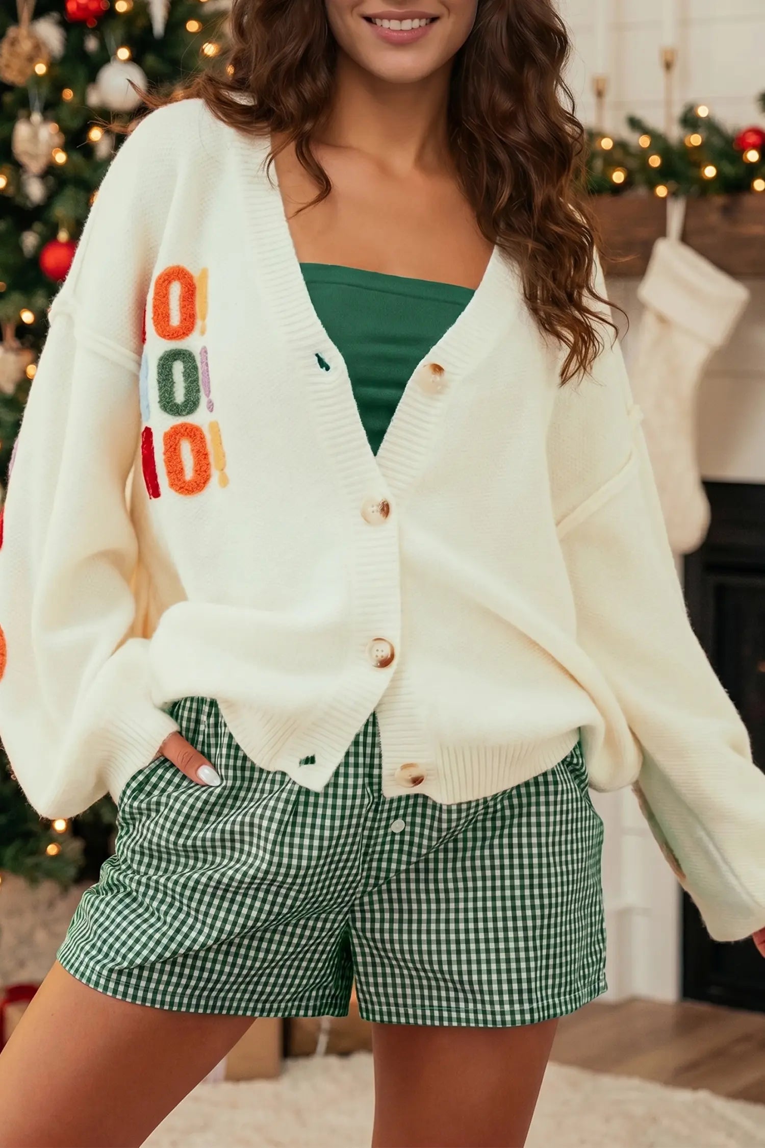 A woman wearing a cream knit cardigan with colorful 'COOL' lettering on the front, styled over a green top and green-and-white gingham shorts in a holiday home setting.