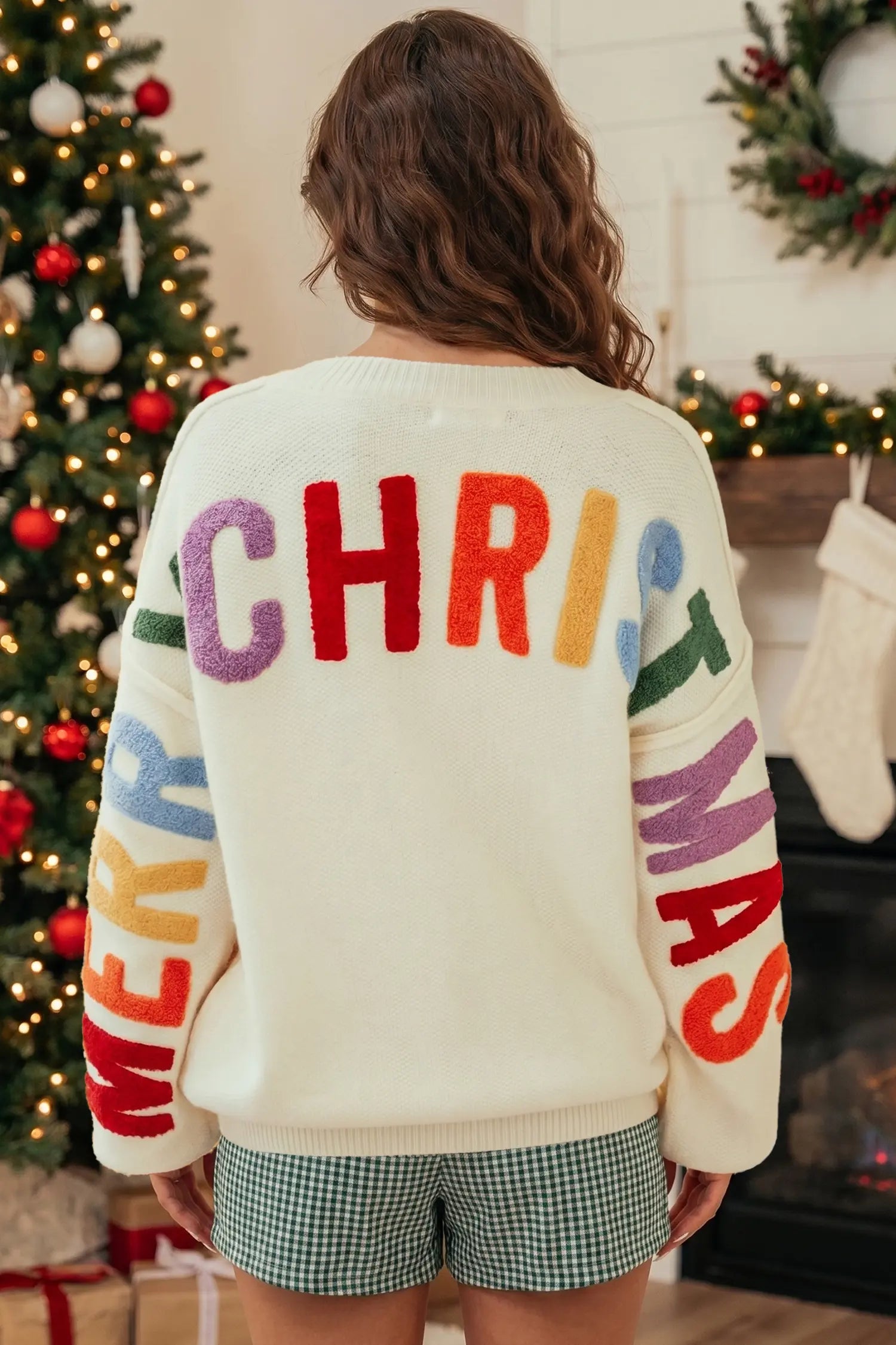 A woman viewed from behind wearing a cream-colored Christmas sweater with colorful fuzzy lettering that reads 'MERRY CHRISTMAS' on the back and sleeves, paired with green and white gingham shorts in a holiday decorated room.