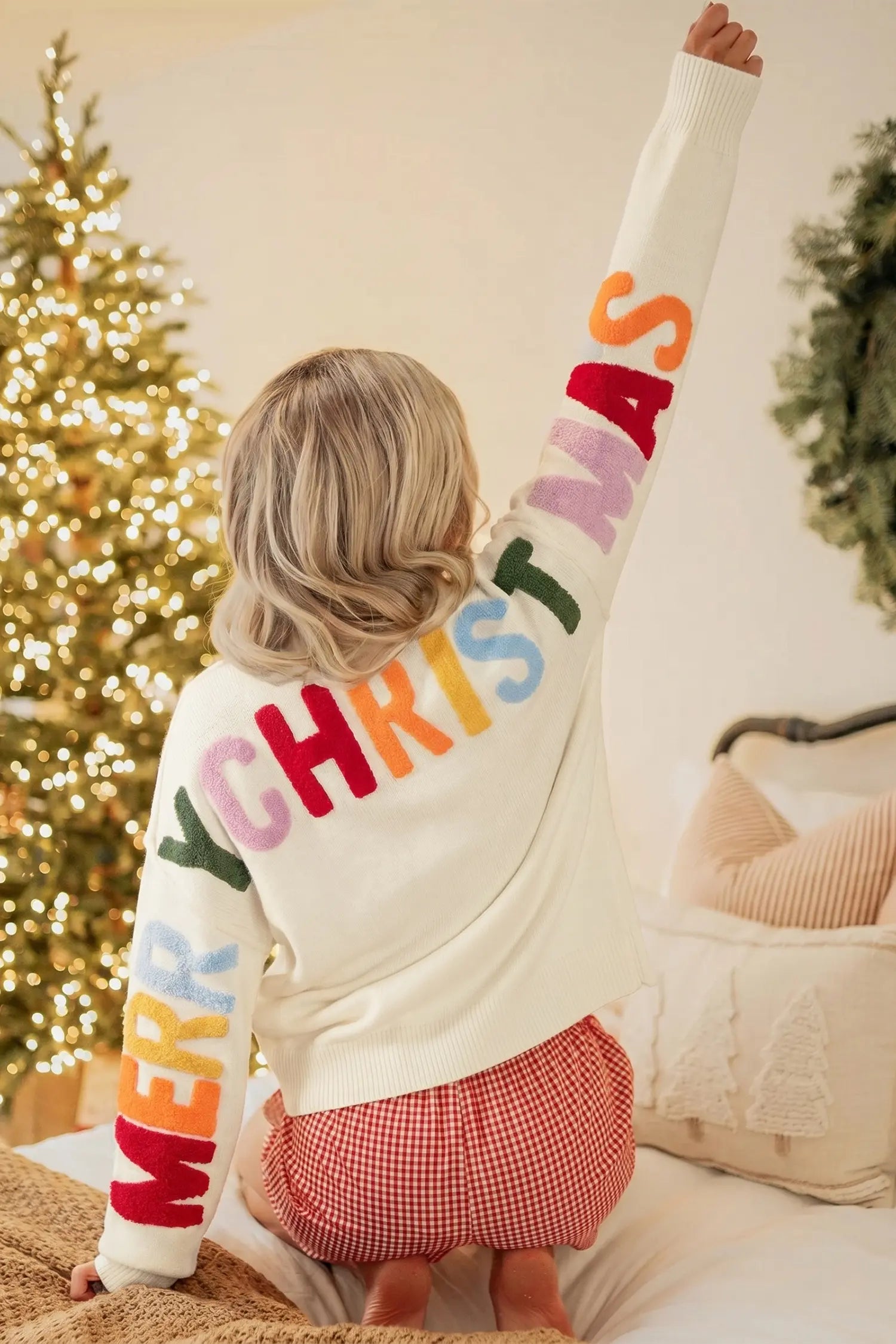A child seen from behind wearing a cream knit sweater with colorful 'MERRY CHRISTMAS' lettering, sitting on a bed in a cozy bedroom with a lit Christmas tree in the background.