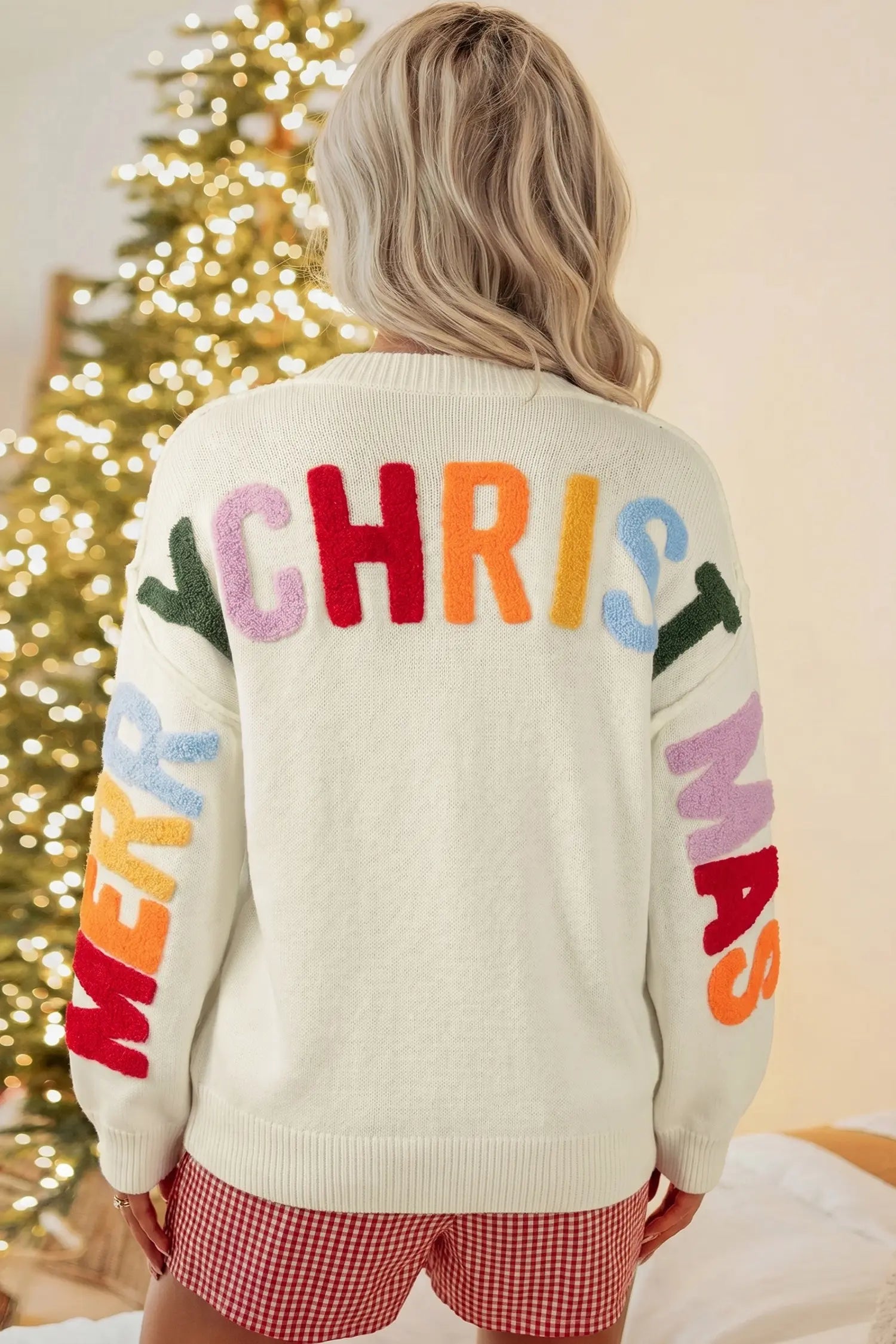 A woman viewed from behind wearing a cream knit sweater with large colorful 'CHRISTMAS' lettering across the back and 'MERRY' on one sleeve, paired with red-and-white gingham shorts in front of a lit Christmas tree.