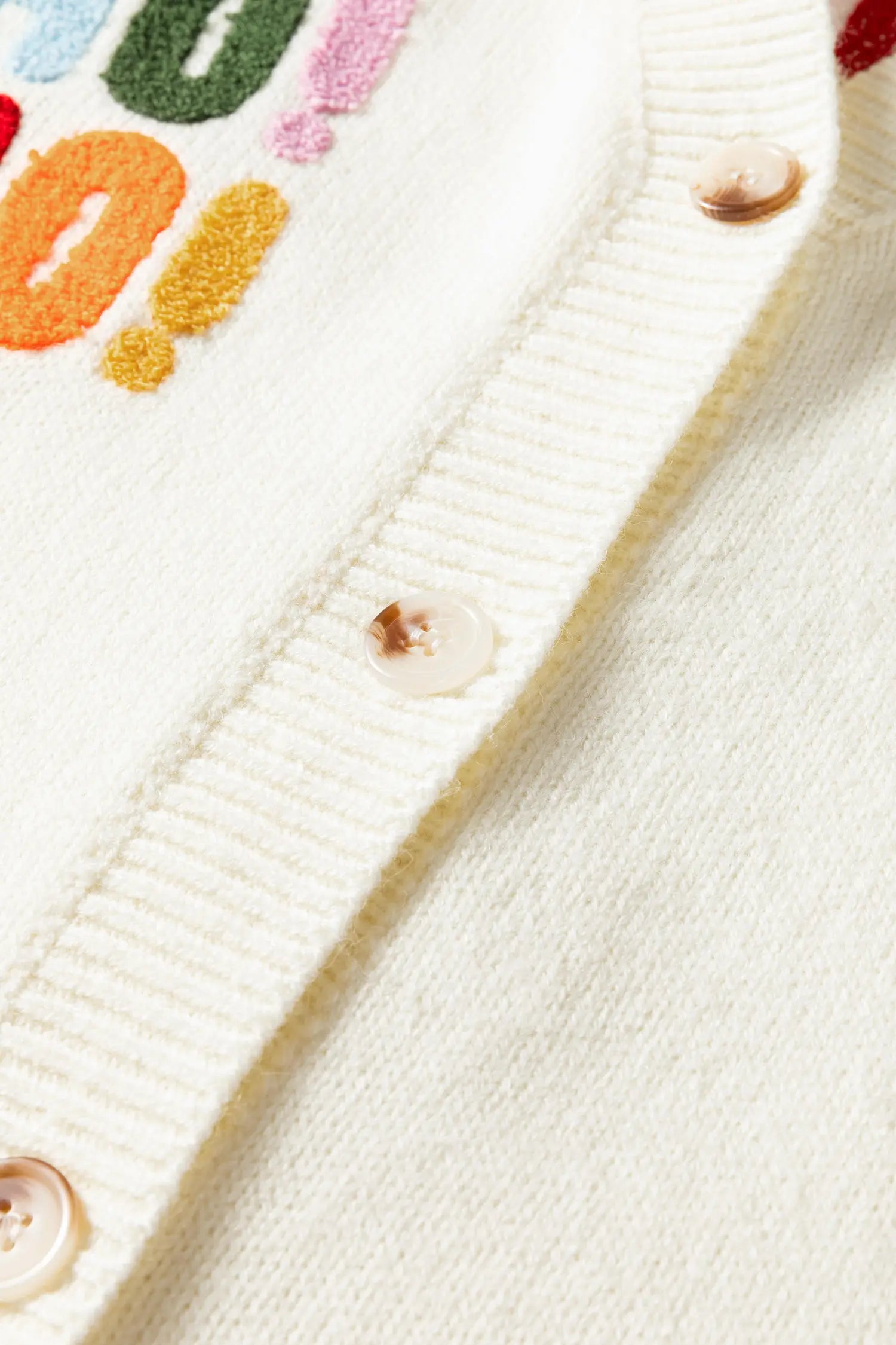 Close-up of a cream-colored knit cardigan featuring colorful embroidered lettering and exclamation marks, ribbed trim, and translucent buttons.