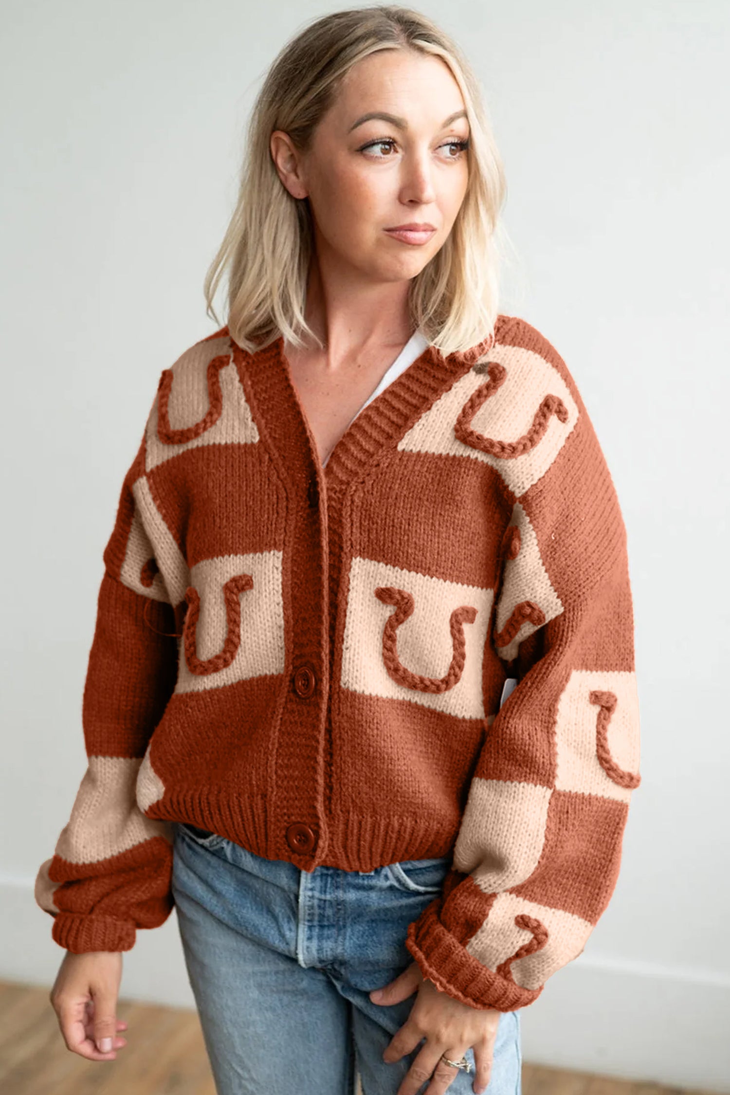 Woman in rust-cream horseshoe cardigan paired with blue jeans against light gray wall.