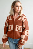 Woman in rust-cream horseshoe cardigan paired with blue jeans against light gray wall.