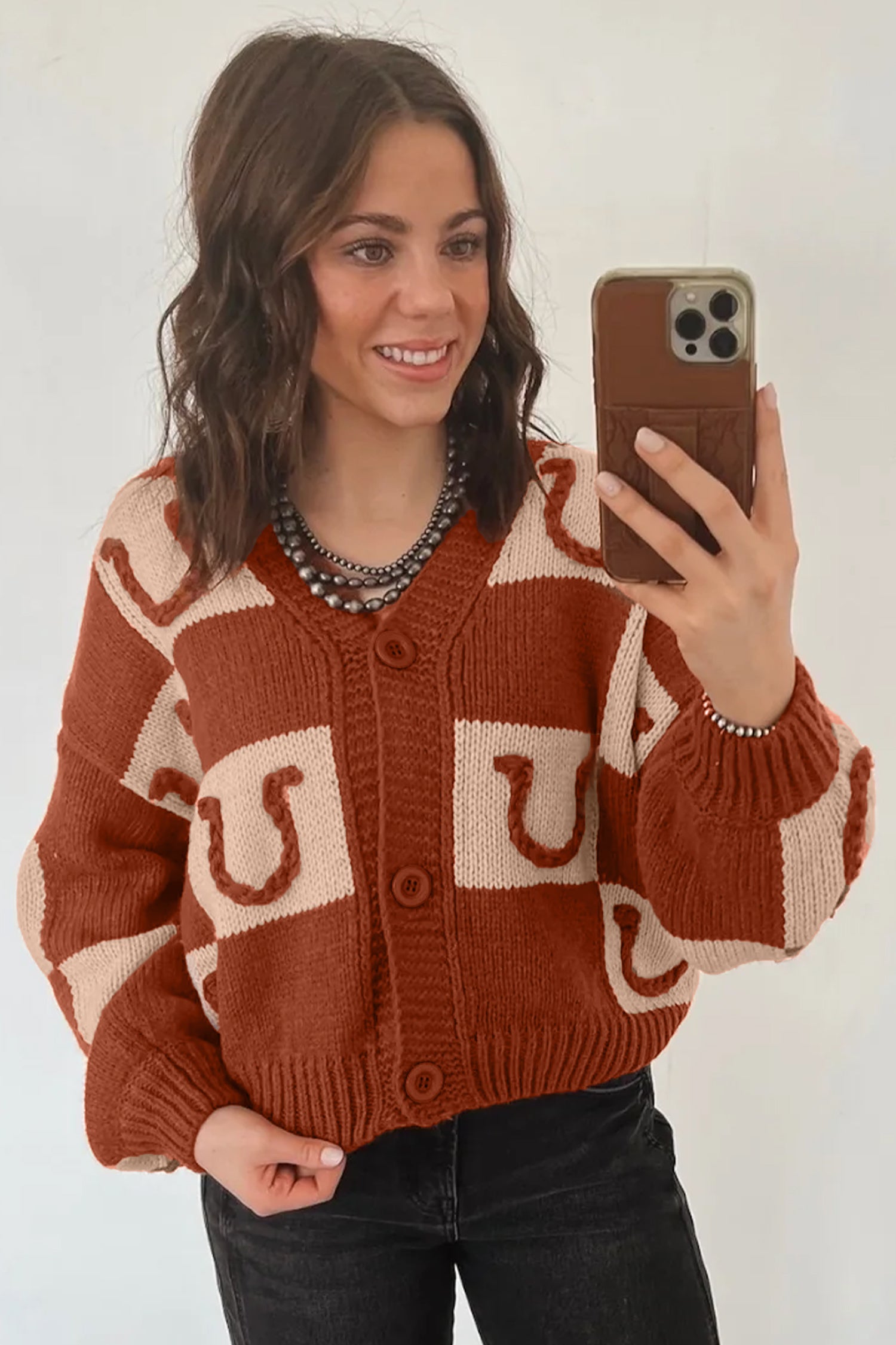 Woman in rust-orange and cream horseshoe cardigan with black jeans and beaded necklace.