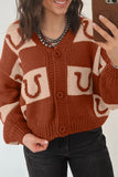 Woman in rust-brown and cream horseshoe cardigan with black jeans and silver necklaces.