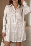 A woman wearing a long-sleeve, button-front shirt dress in a shimmering champagne beige fabric, standing in a softly lit indoor setting.