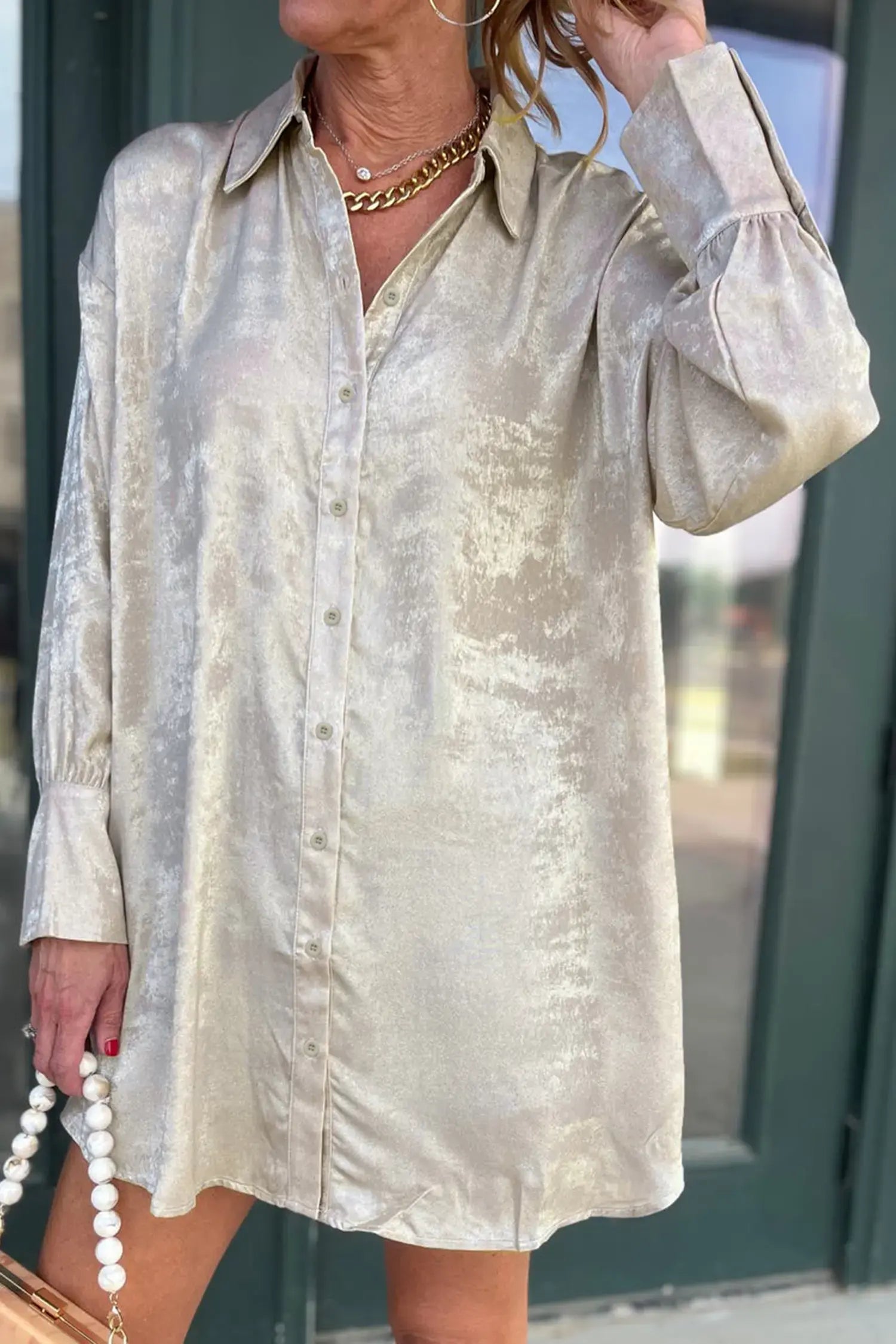 A woman wearing a long-sleeve, button-front shirt dress in a champagne-colored crushed satin fabric, standing in front of a dark green door.
