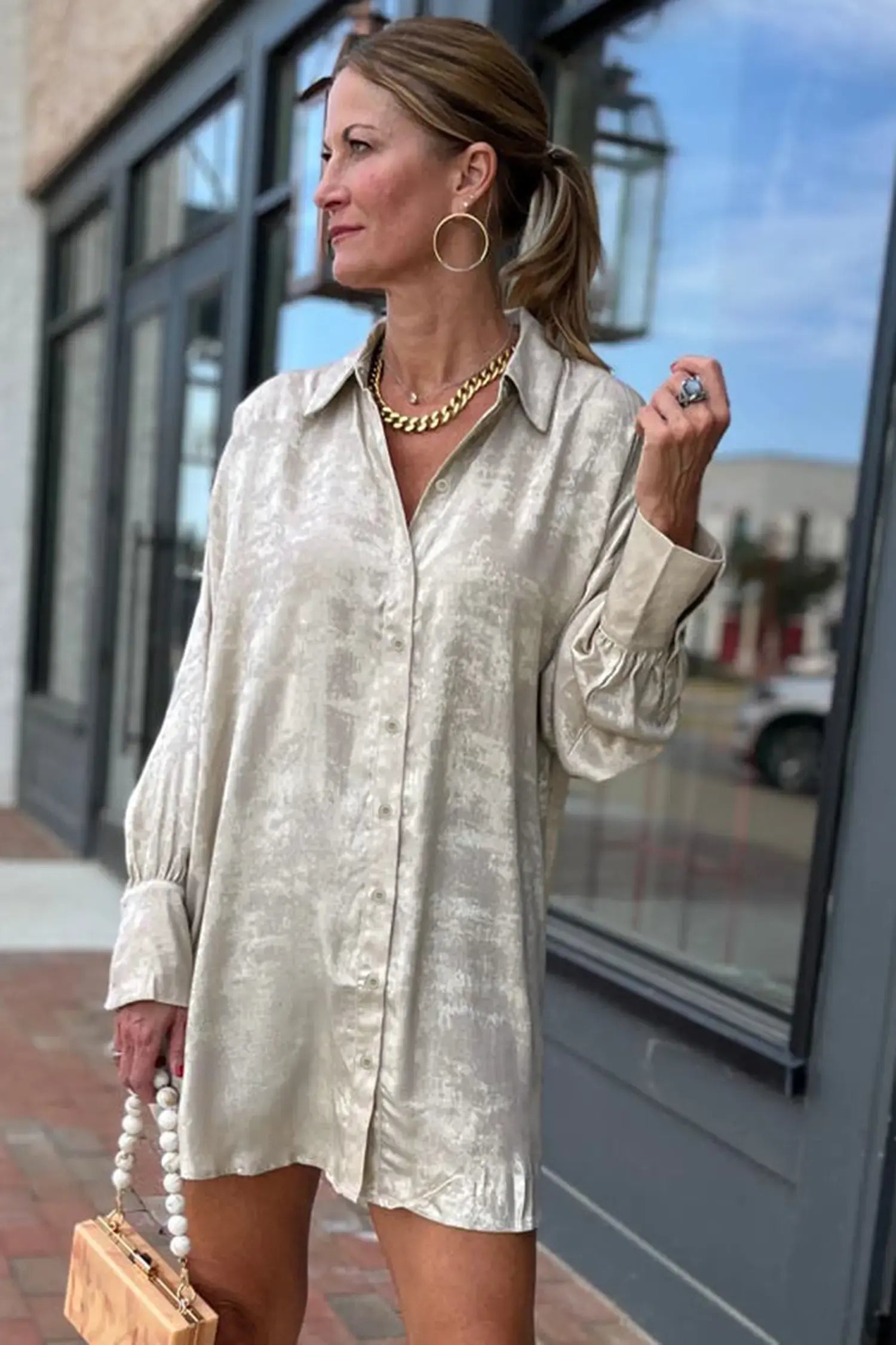 A woman wearing a light beige, textured long-sleeve button-up shirt dress, standing outdoors in front of a storefront window.