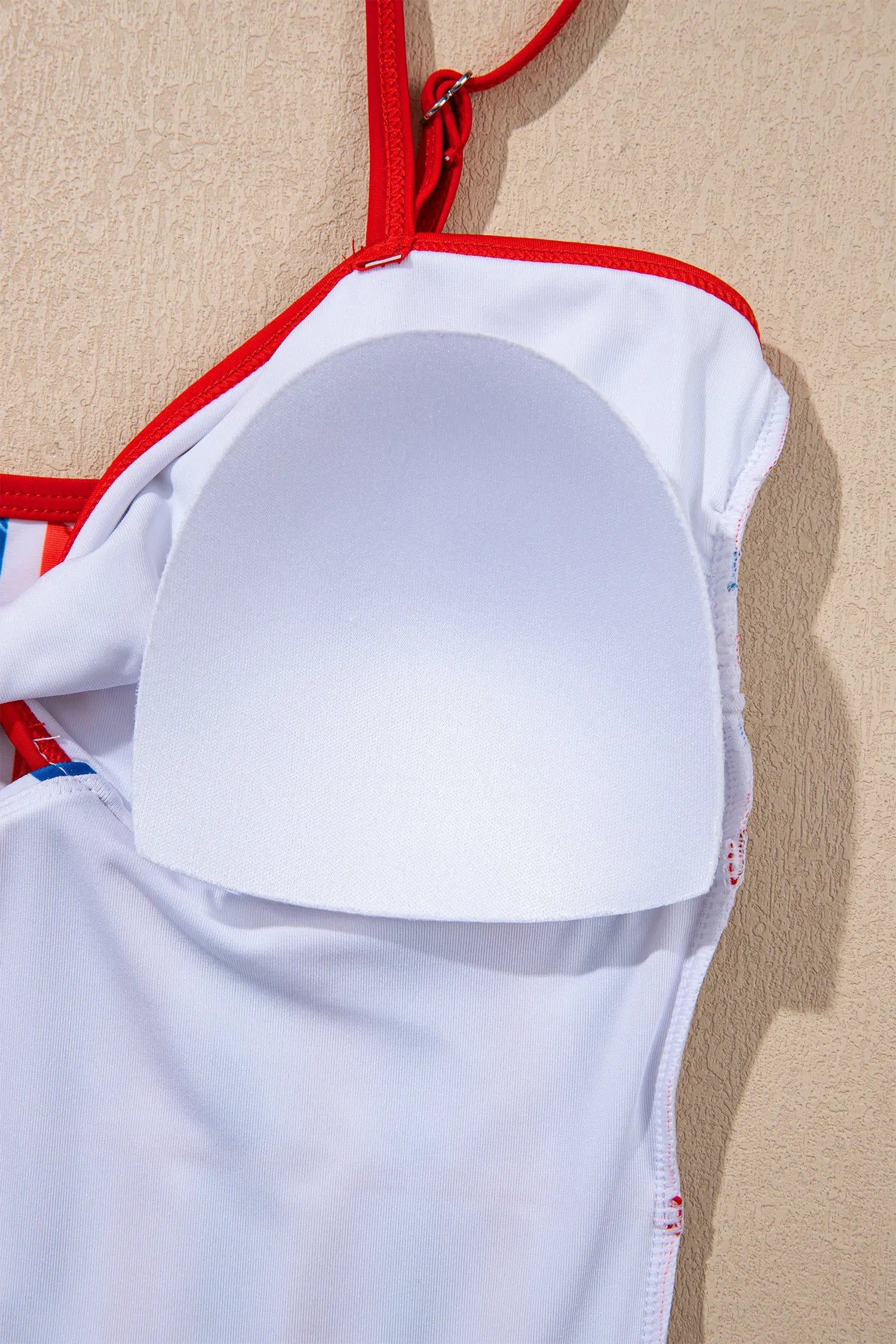 Close-up of a white strapless bra or swimsuit top with red trim and a white padded underband, shown against a beige textured wall.