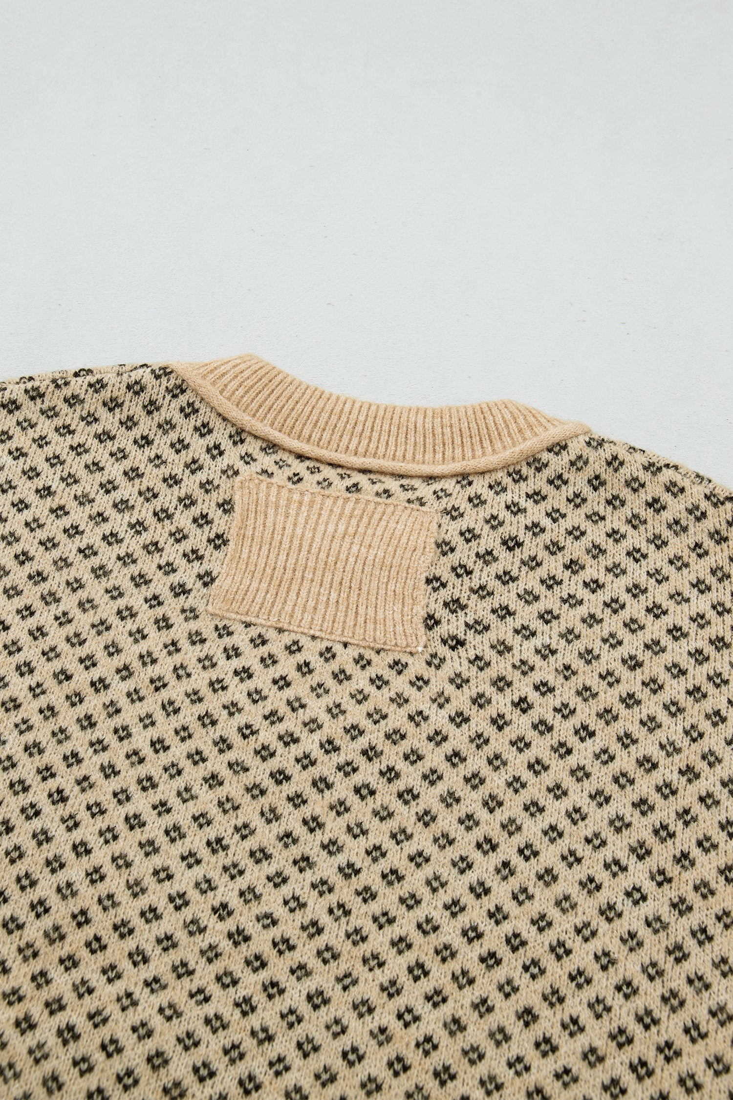 Close-up of the back of a knit sweater with a beige ribbed crew neck and a small rectangular patch on the upper back, over a black and beige micro-pattern.