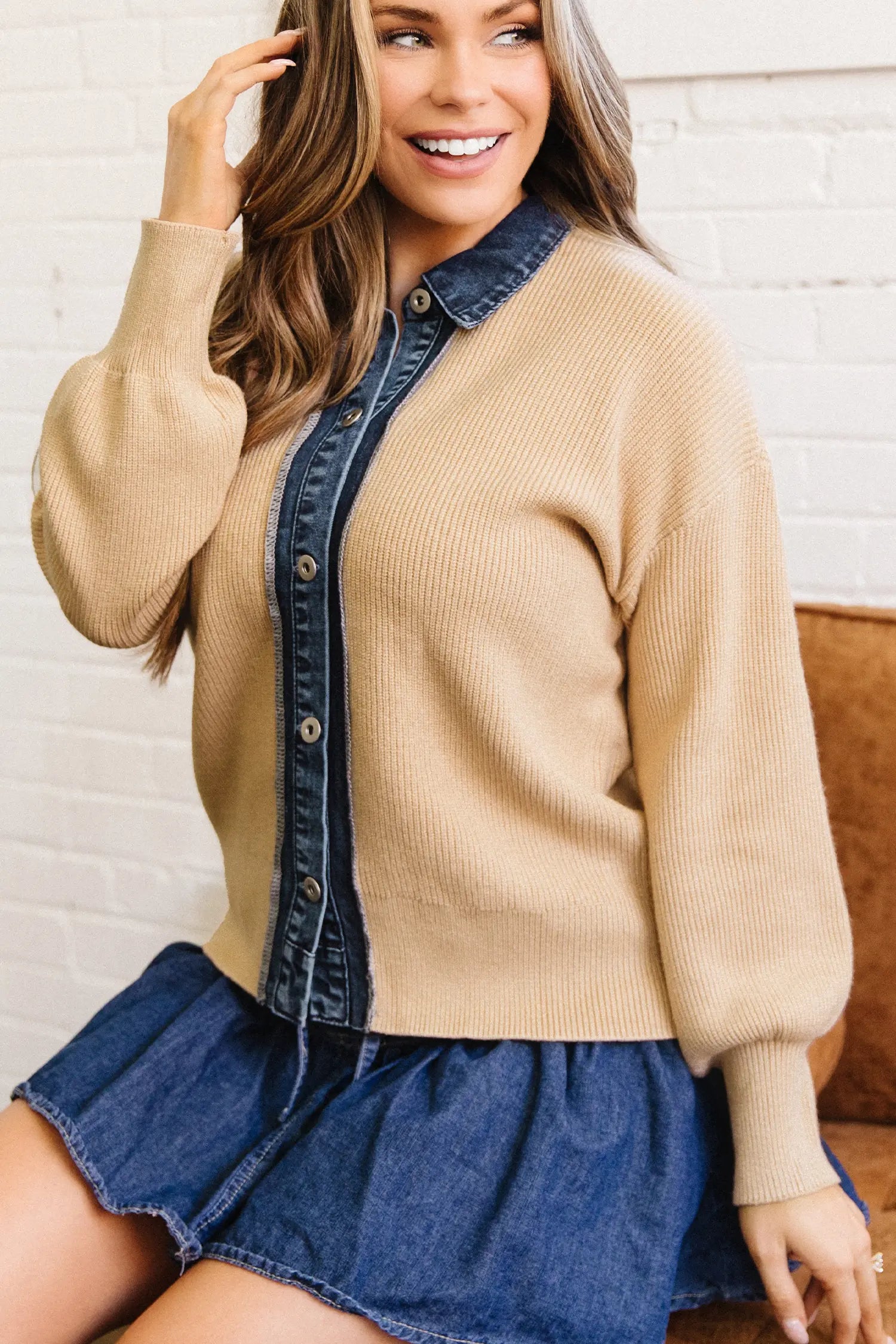 A woman wearing a beige ribbed knit cardigan with a dark blue denim button-front placket and collar, paired with a denim mini skirt, posed in front of a white brick wall.