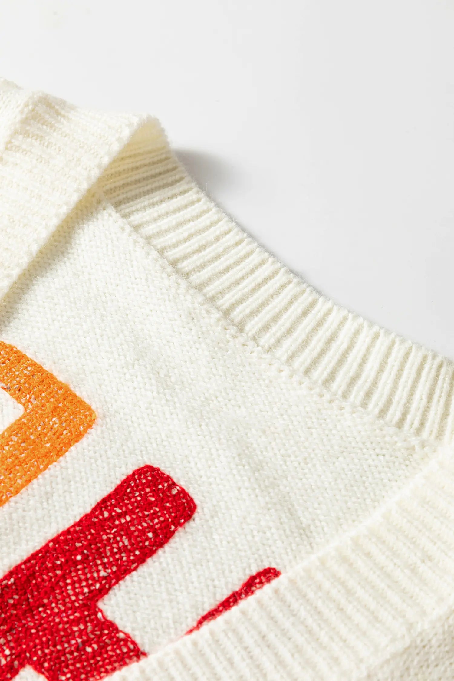 Close-up of a cream-colored knit sweater showing the ribbed crew neck and a portion of a colorful graphic with orange and red lettering.