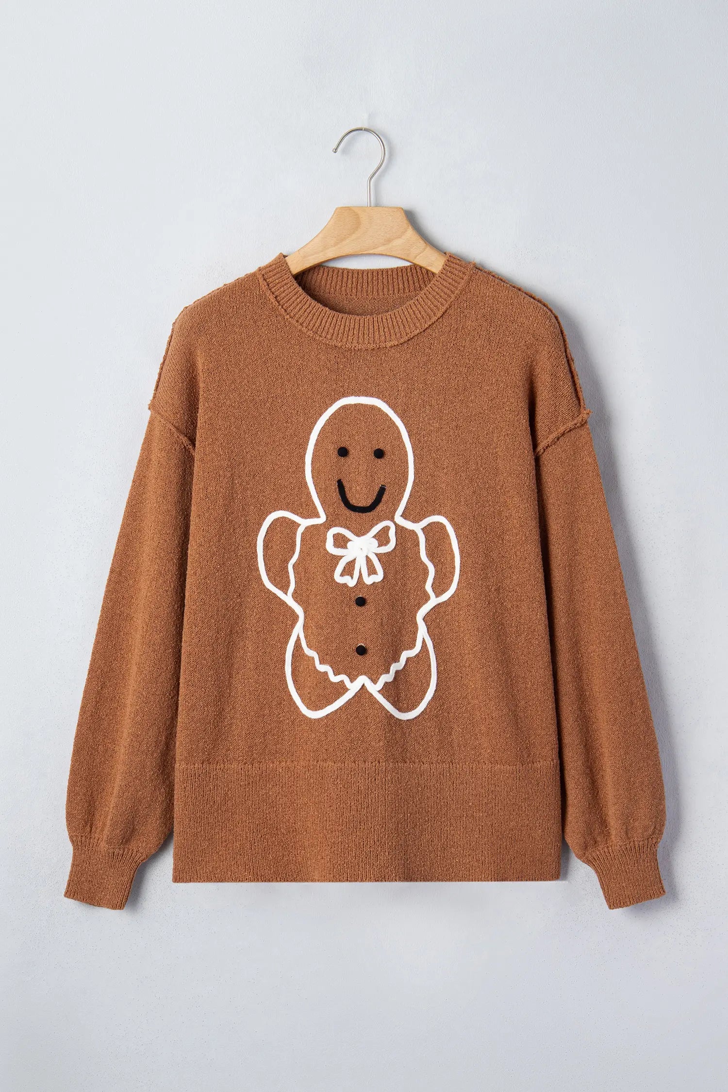 A brown knit crewneck sweater with a white and brown gingerbread man graphic on the front, hanging on a wooden hanger against a light gray background.