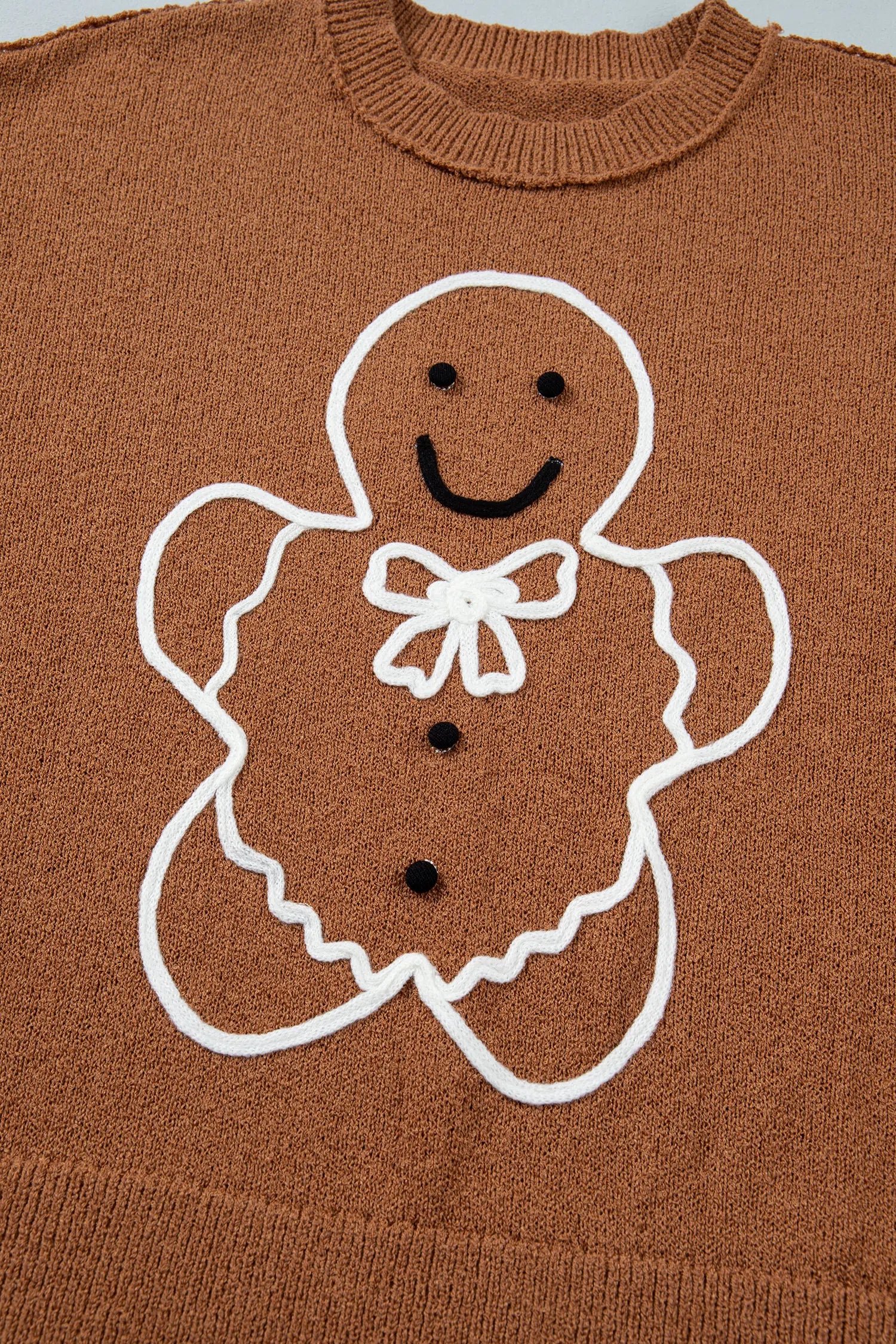 A close-up of a brown knit sweater featuring a white-outlined gingerbread man design with black embroidered facial features and a white bow on the chest.