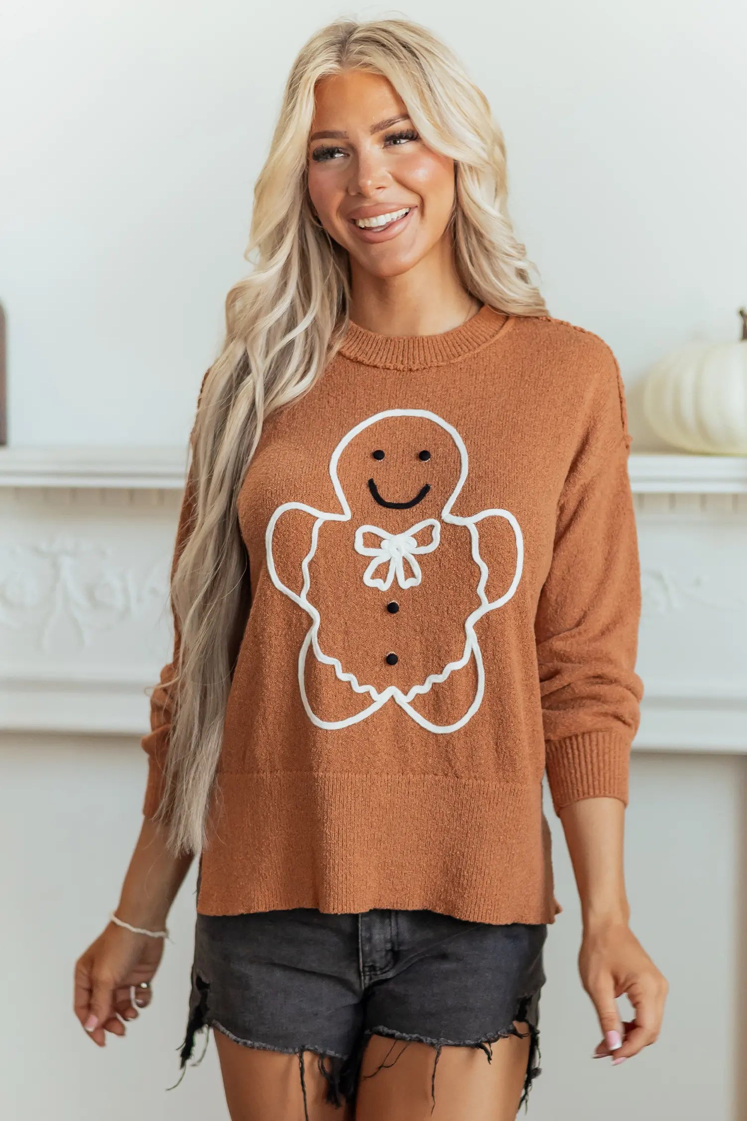 A woman wearing a brown crewneck sweater with a white gingerbread man graphic on the front, paired with dark denim shorts, standing in front of a white fireplace mantel.