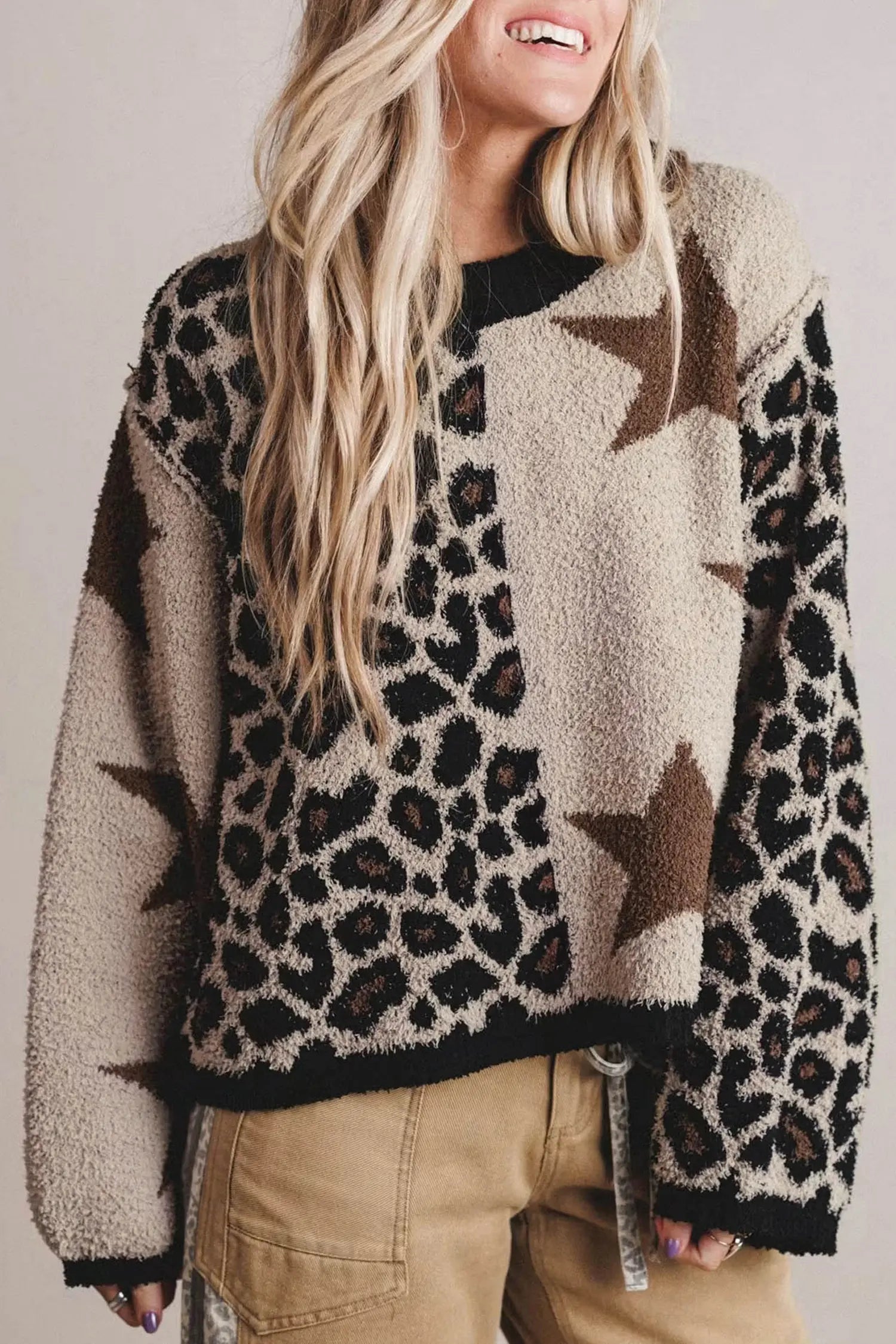 A woman wearing a fuzzy knit sweater with a leopard print and star pattern in beige, brown, and black, paired with tan pants against a neutral studio background.