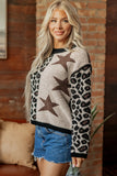 A blonde woman wearing a fuzzy knit sweater with a beige and black leopard print pattern, large brown star motifs on the front, and leopard-print sleeves, paired with denim shorts, standing in front of a brick wall.