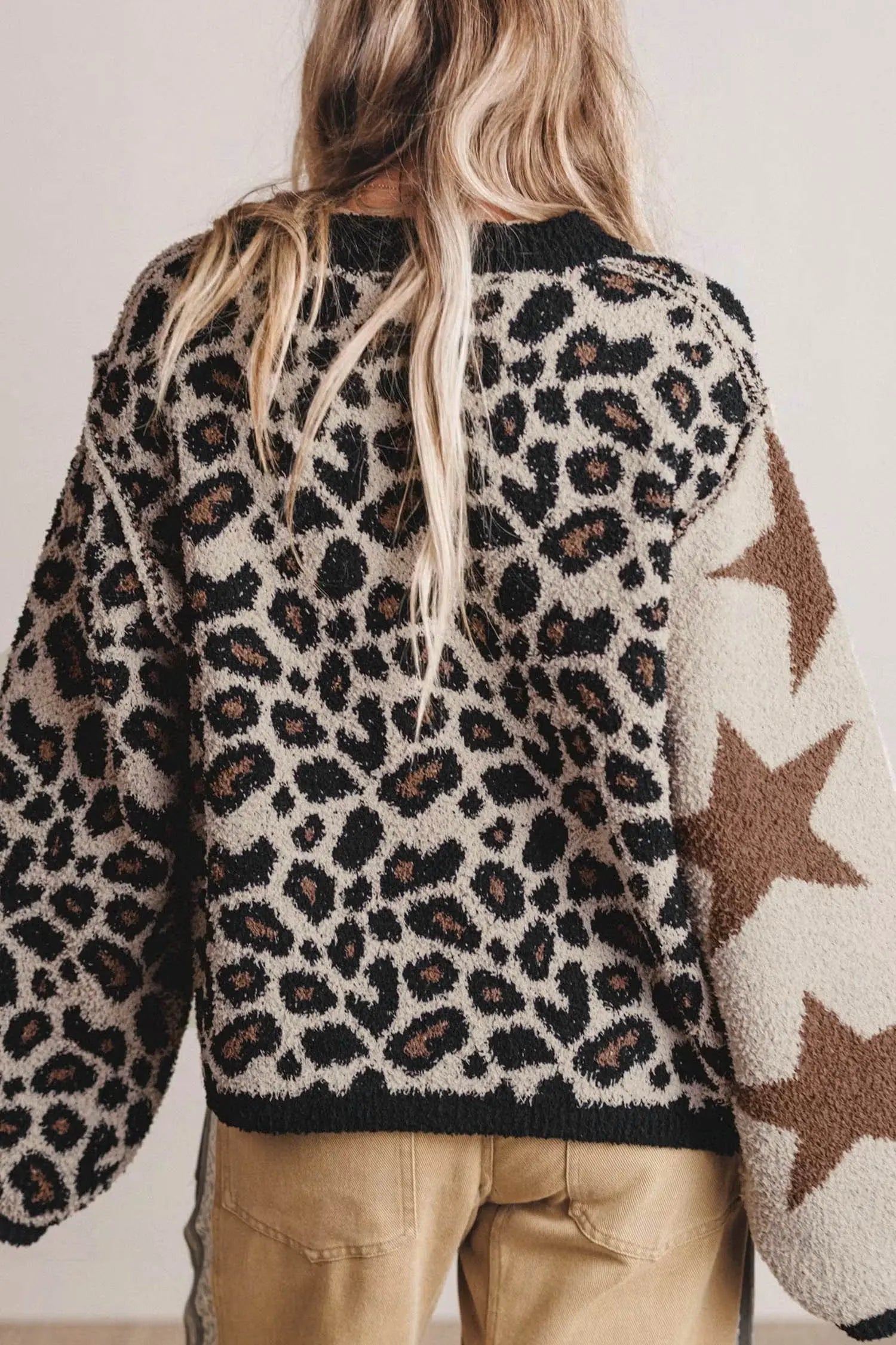 A person shown from the back wearing a fuzzy knit sweater with a leopard print pattern on the main body and large brown stars on one sleeve, paired with tan pants against a plain light background.