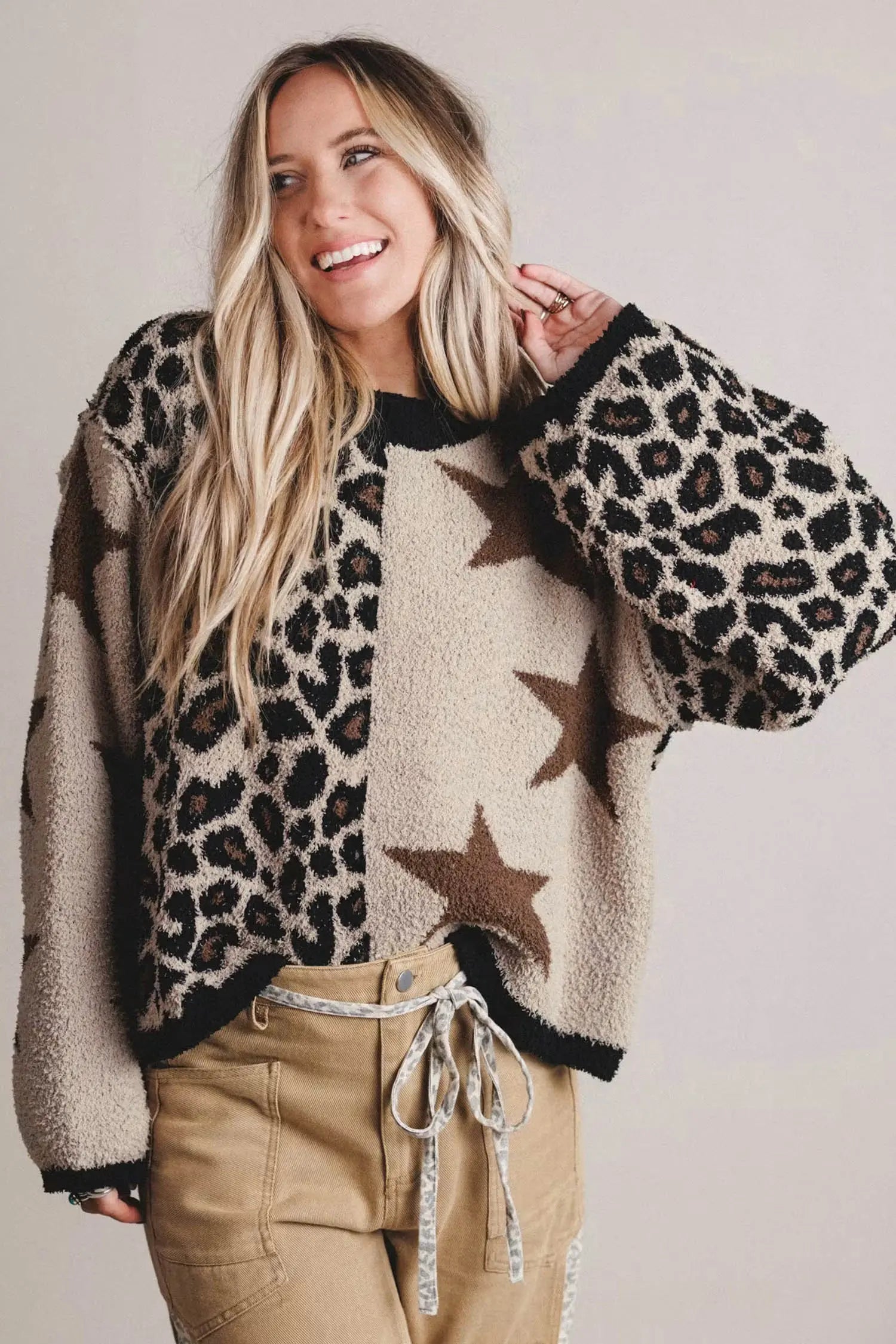 A woman wearing a fuzzy knit sweater with a mixed pattern of leopard print, solid beige, and brown stars, paired with khaki pants, against a plain light gray background.