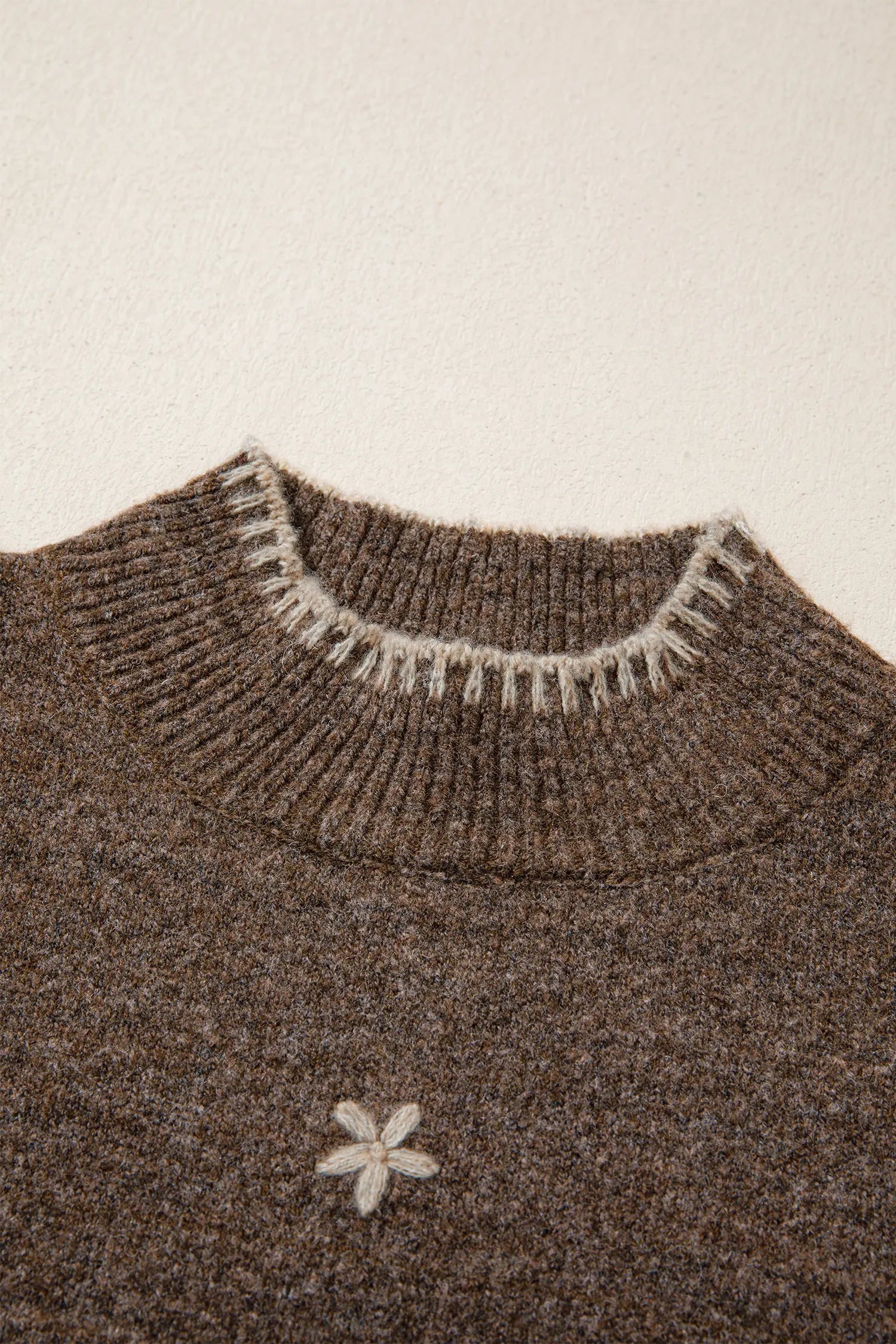 Close-up of a brown knit sweater with a ribbed crew neck featuring white fringe trim and a small white embroidered flower on the chest.