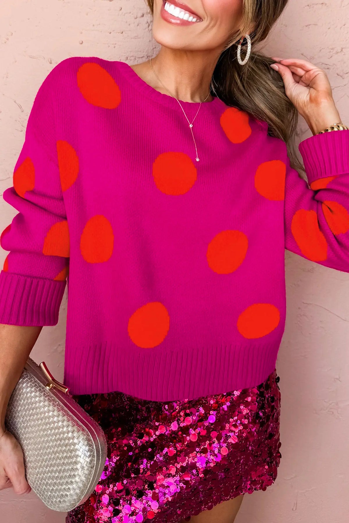 A woman wearing a bright pink knit sweater with large orange-red polka dots, paired with a shiny magenta sequined skirt and holding a silver clutch against a pink wall.
