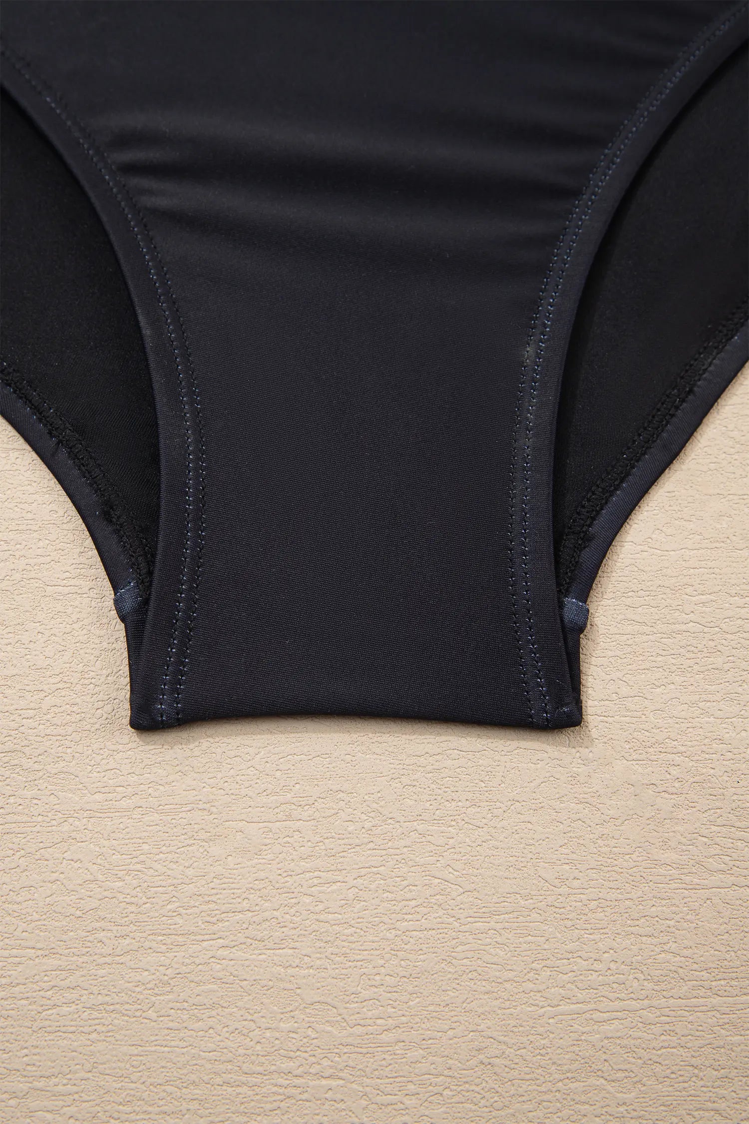 Close-up of a black bikini bottom or underwear leg opening laid flat on a light beige textured surface.
