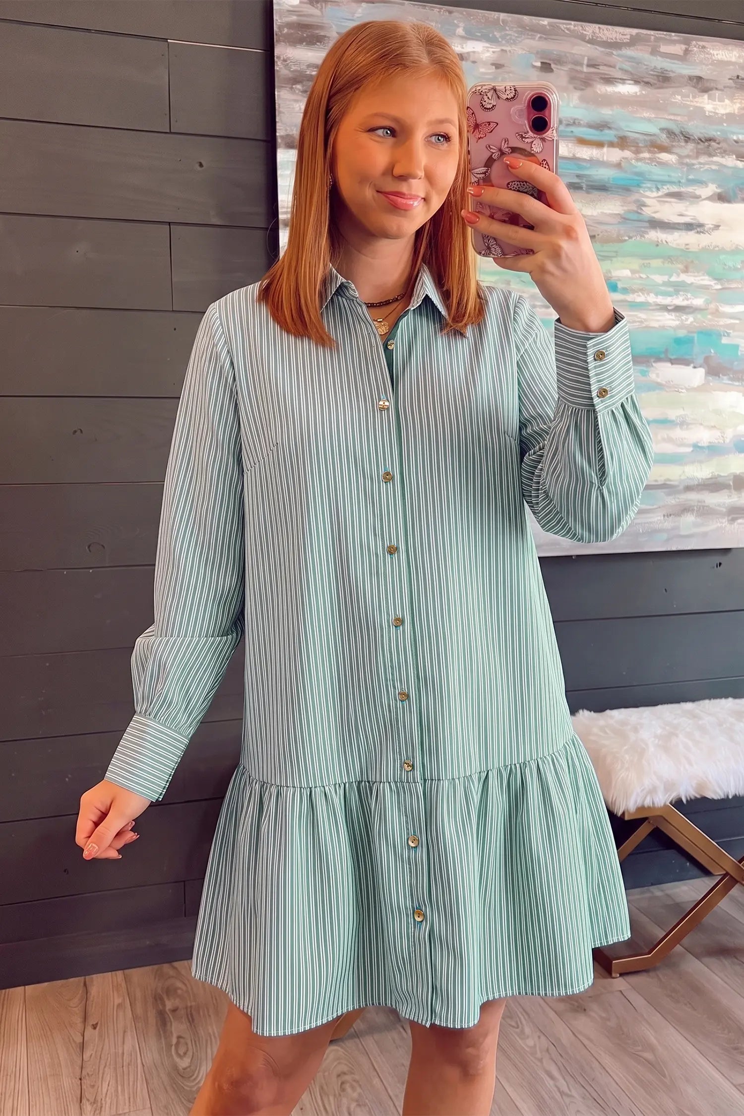 A woman takes a mirror selfie wearing a mint green and white vertically striped long-sleeve button-down shirt dress with a ruffled hem, set against a dark wood-paneled wall.
