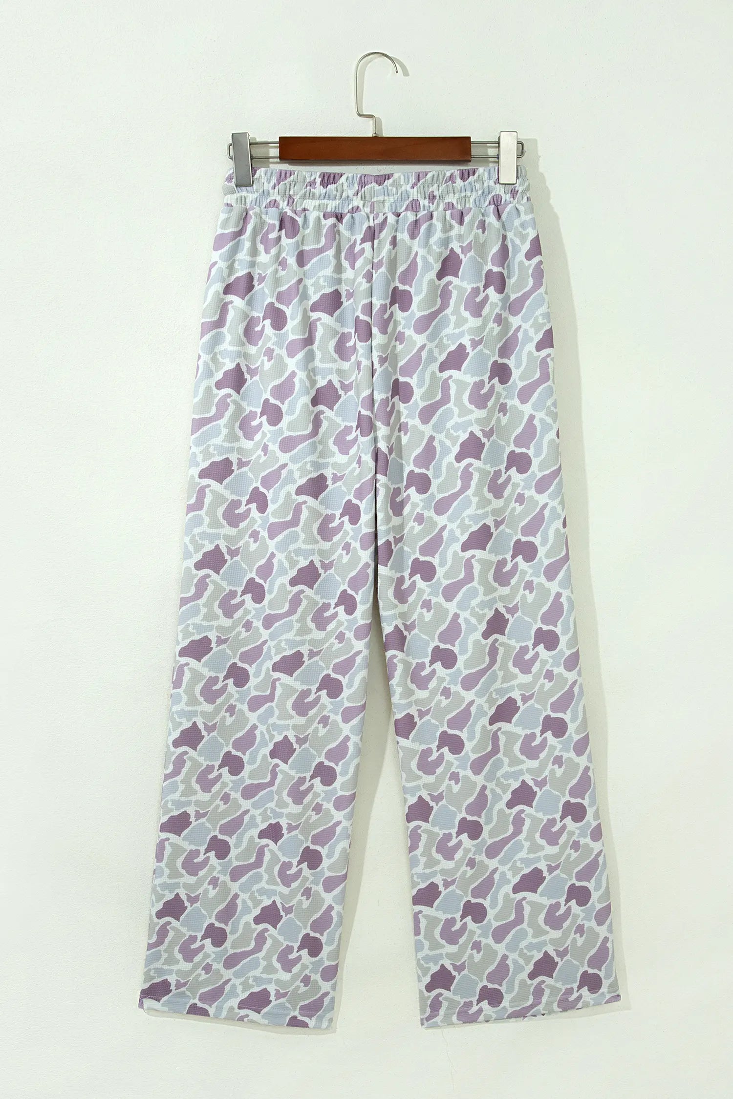 A pair of wide-leg pants with an all-over abstract pattern in purple, gray, and white, hanging from a wooden hanger against a plain off-white wall.