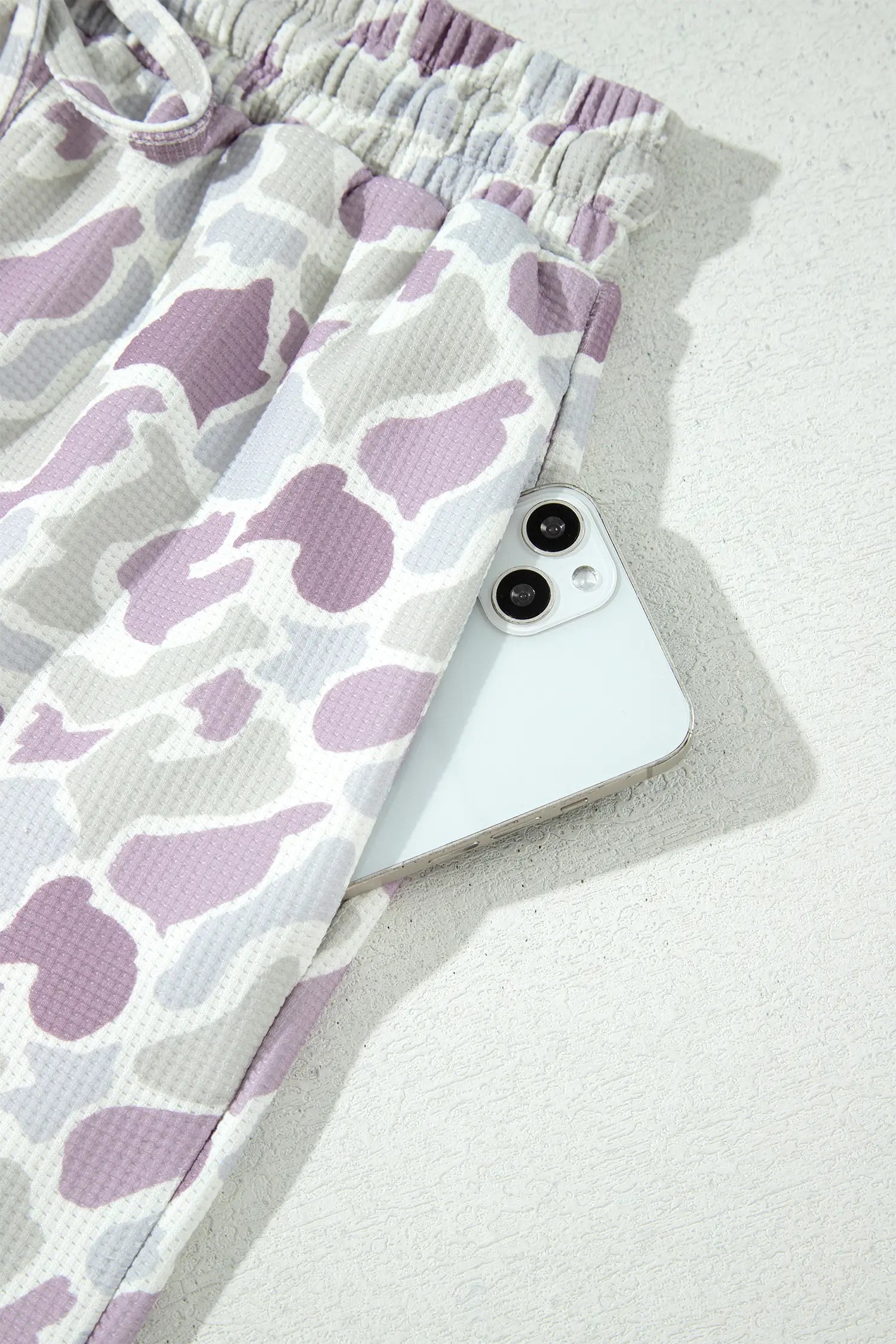 A close-up of patterned lounge pants in a waffle texture, featuring a purple and white abstract cow-print design. A white smartphone is partially tucked under the side of the pants, which are laid out on a textured light gray surface.
