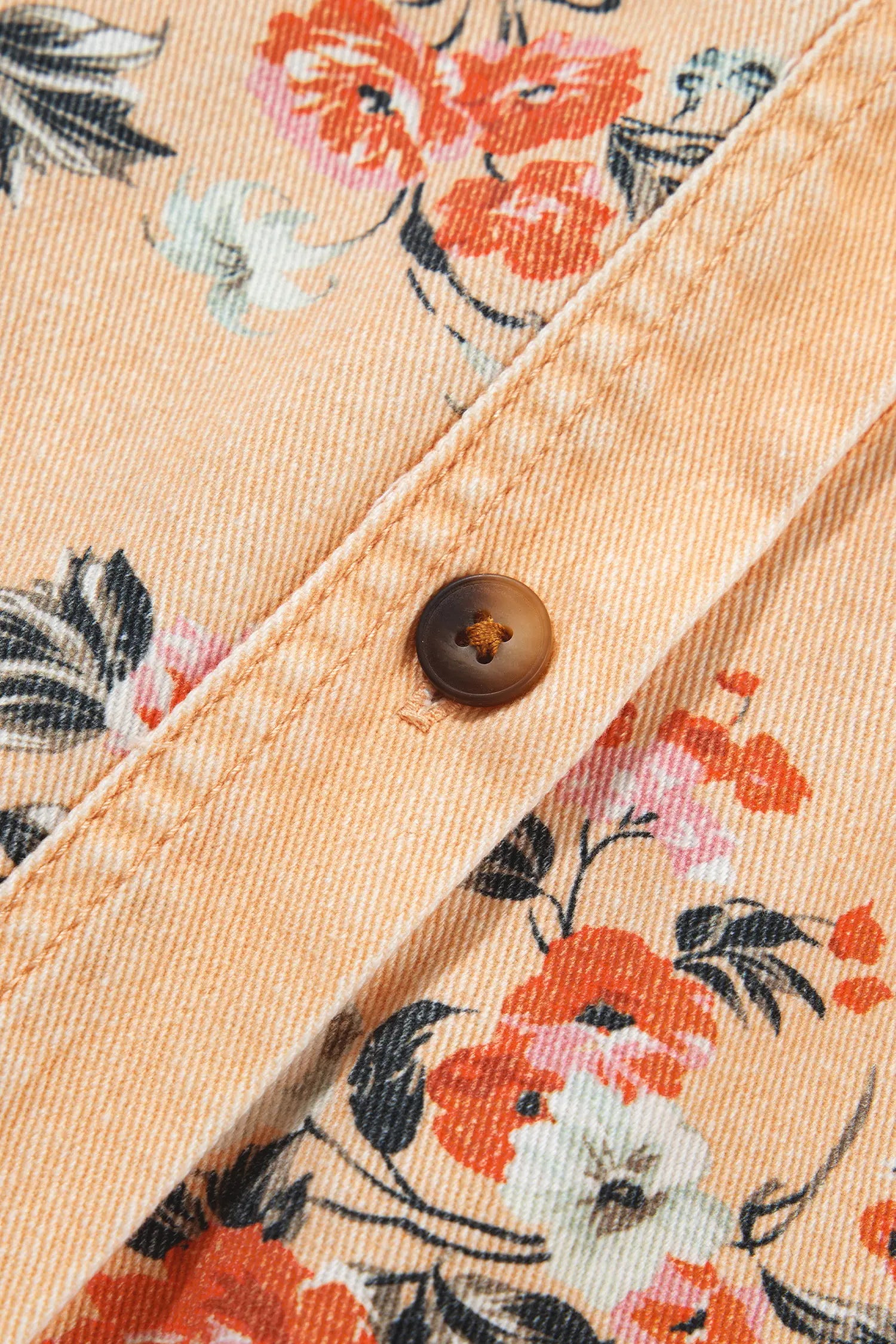 Close-up of peach denim shirt with orange, pink, white floral print, black leaves, and brown button.