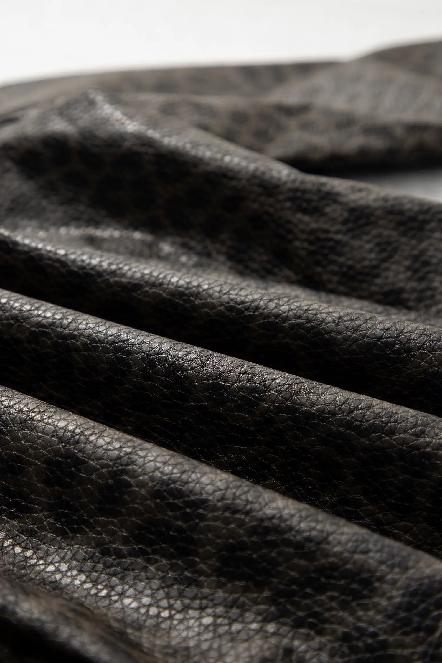 Close-up of dark textured leather with a pebbled grain and soft folds, photographed against a light neutral background.