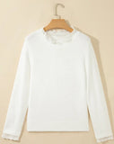 A white long-sleeve knit sweater with delicate lace trim at the crew neckline and cuffs, hanging on a wooden hanger against a warm beige background.